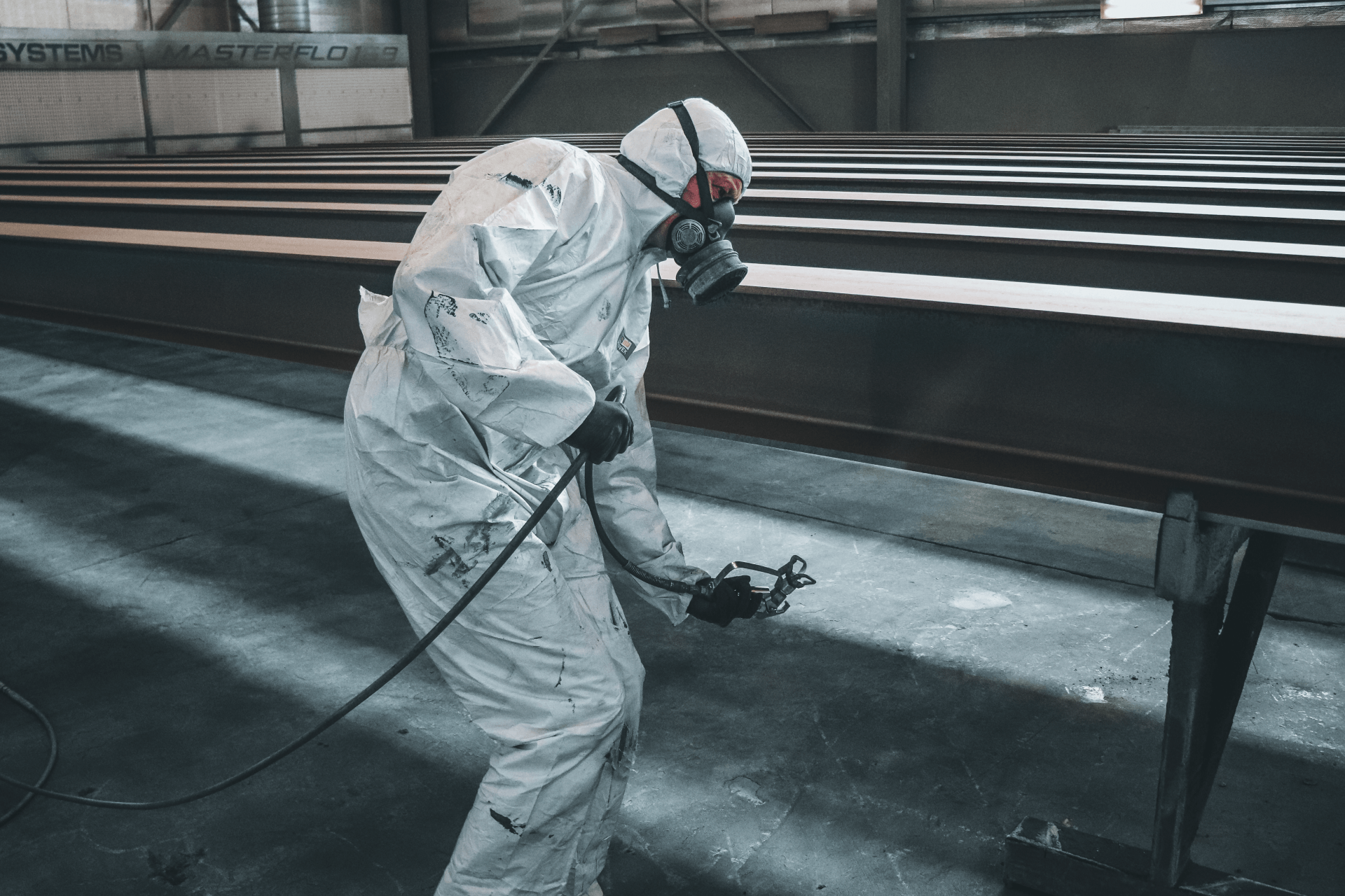 Person in protective suit spraying paint onto a metal beam in an industrial setting.