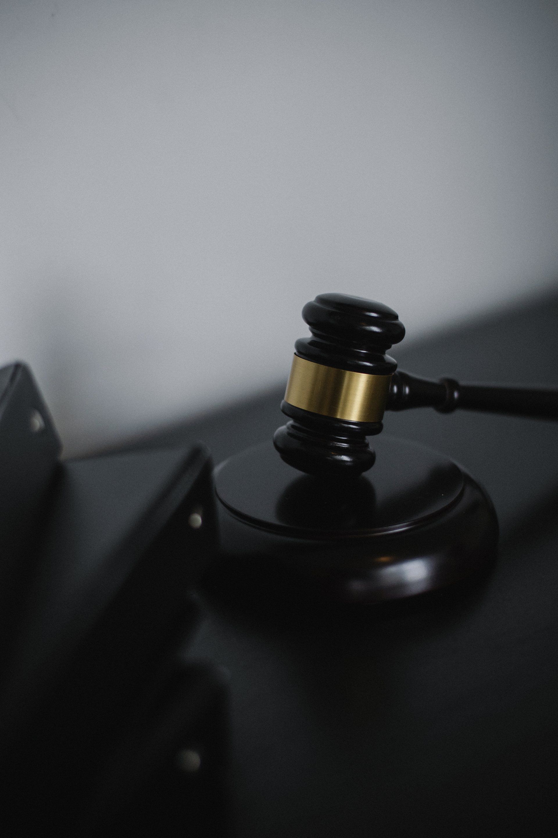 A black wooden gavel with a gold band rests on a matching sounding block against a dark, minimalist background.