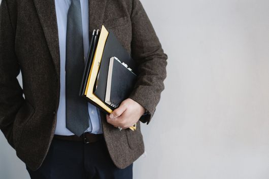 Person in a brown blazer holding folders and a notebook against a gray wall
