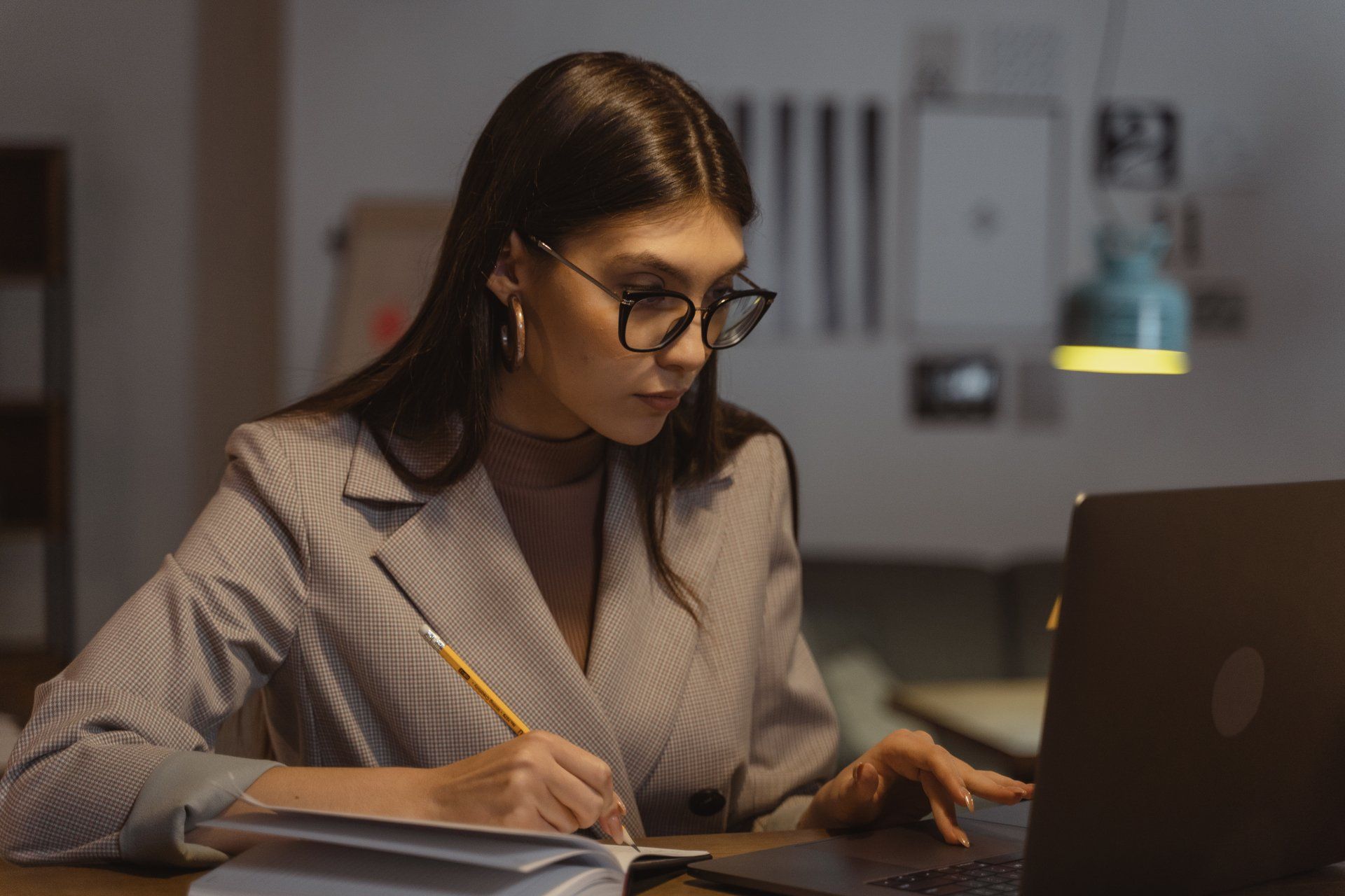 A woman is sitting at a desk using a laptop computer and writing in a notebook.