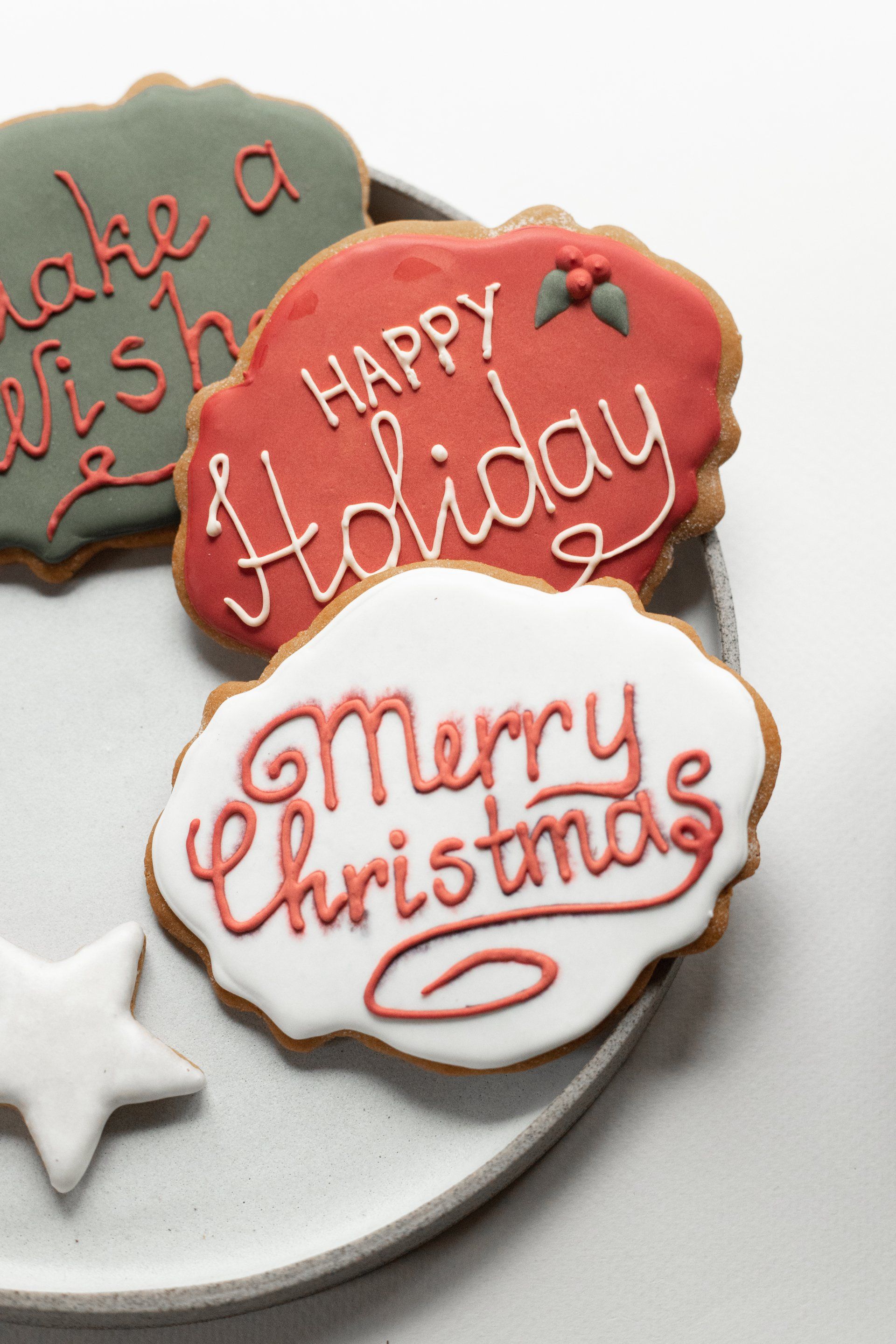 Three decorated Christmas cookies with holiday greetings on a white plate, alongside a small white star-shaped cookie.