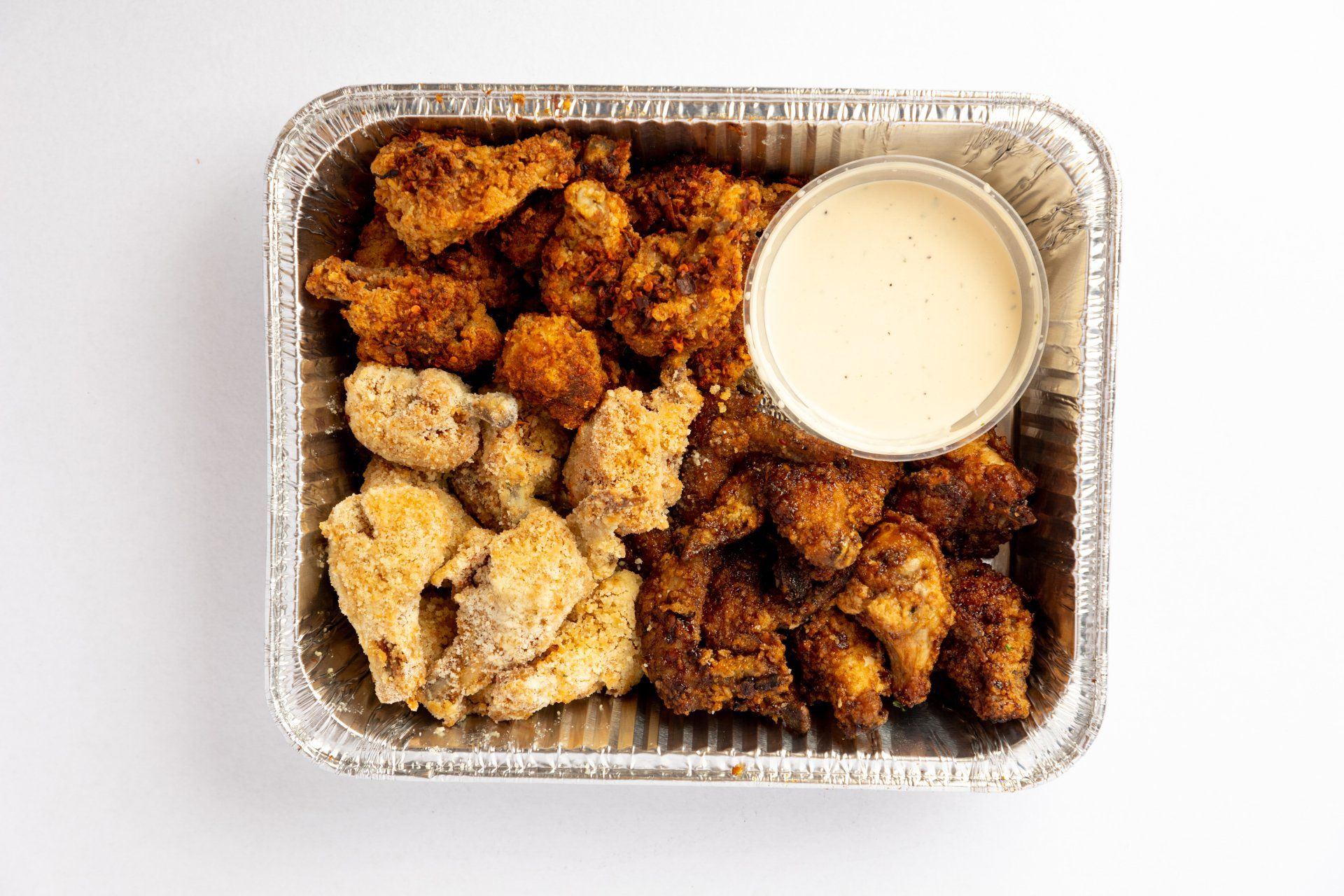 A rectangular foil container filled with a mix of fried chicken wings and a side cup of creamy white dipping sauce.