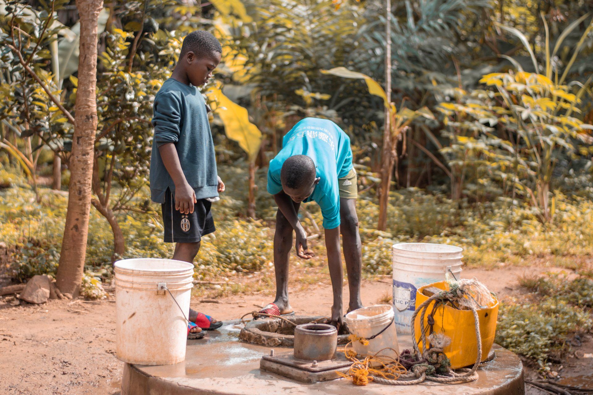 Two boys working near a well, one bent over, surrounded by buckets, outdoors.