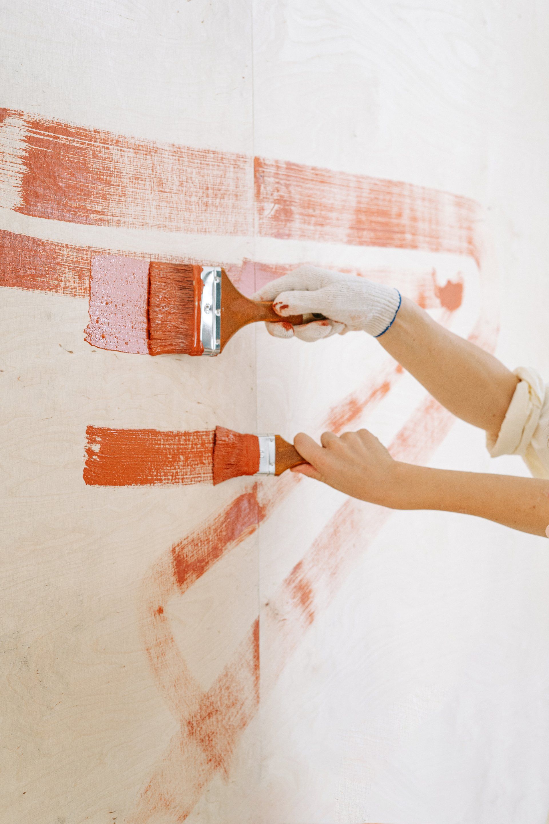 Two hands painting a white wall with orange paint using paintbrushes.