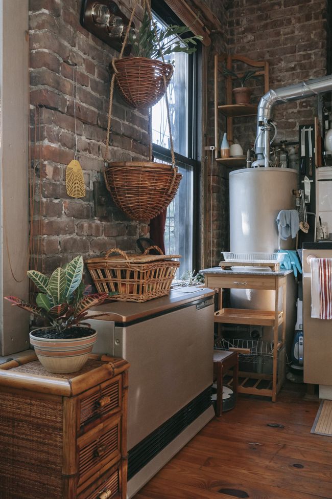 A corner of a room with exposed brick walls, a plant on a woven chest, hanging baskets, and a metal water heater.