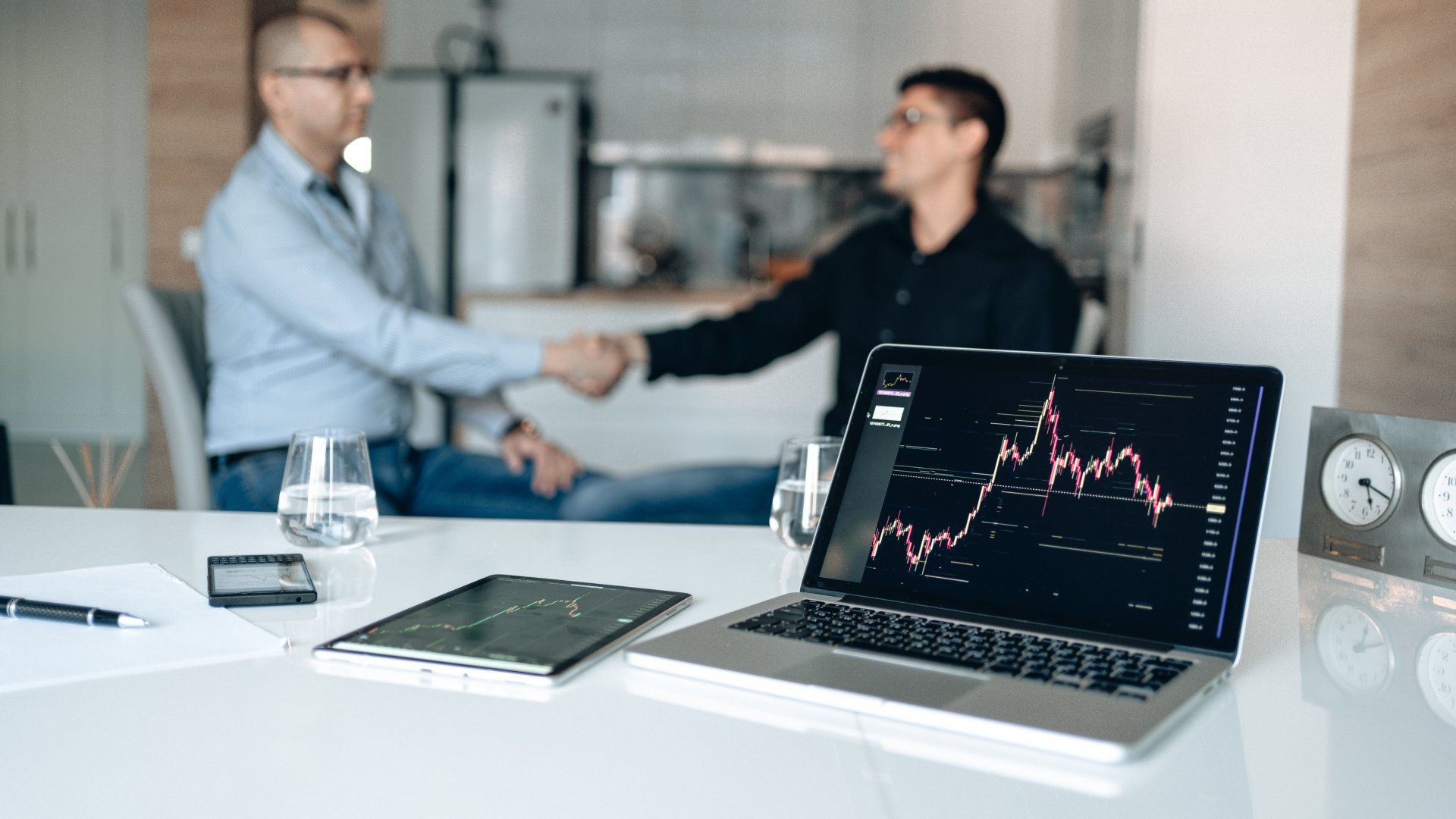 Two professionals shaking hands in the background with a laptop displaying an upward-trending graph Two professionals shaking hands in the background with a laptop displaying an upward-trending graph