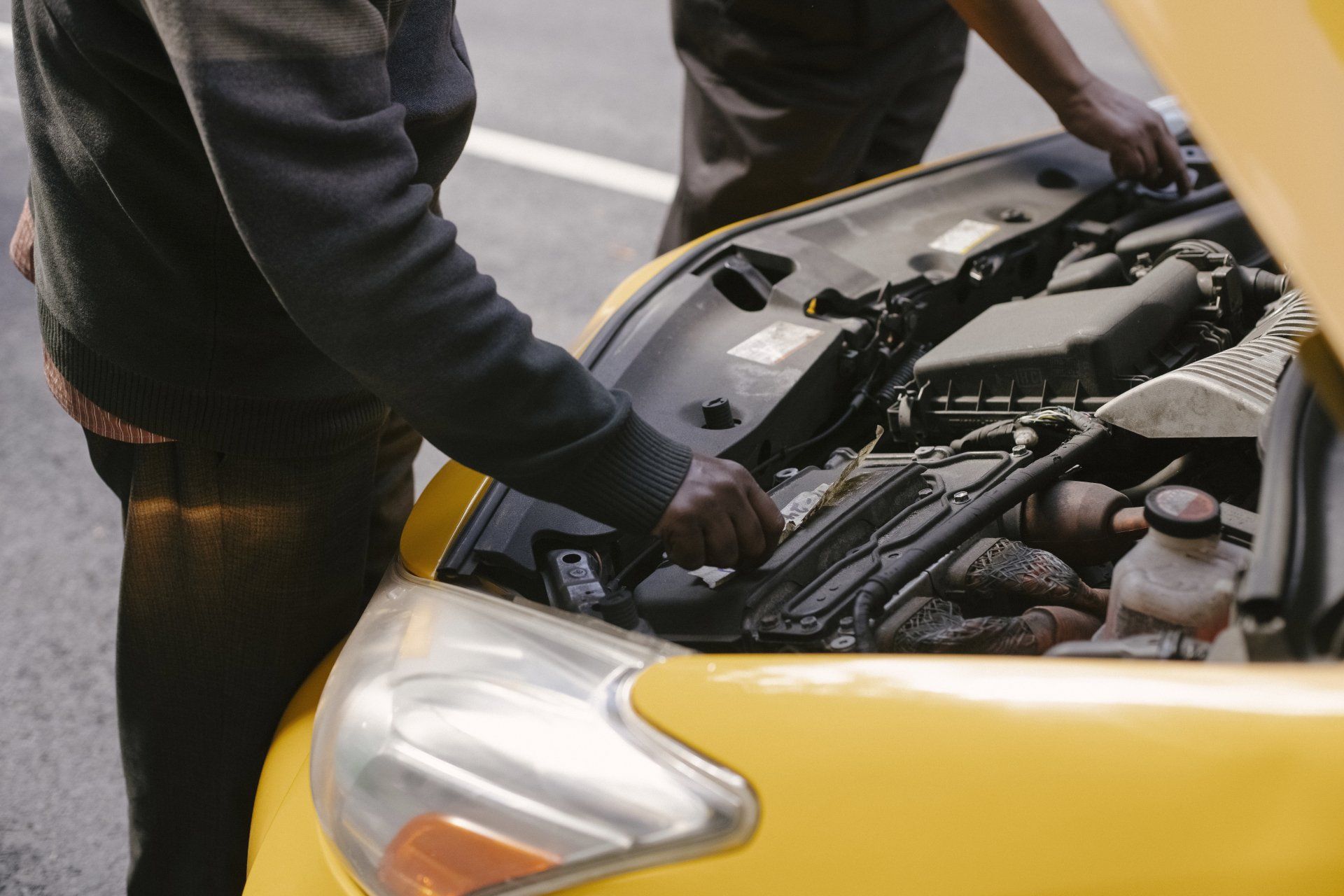 Two people looking under the hood of a yellow car in an outdoor setting.