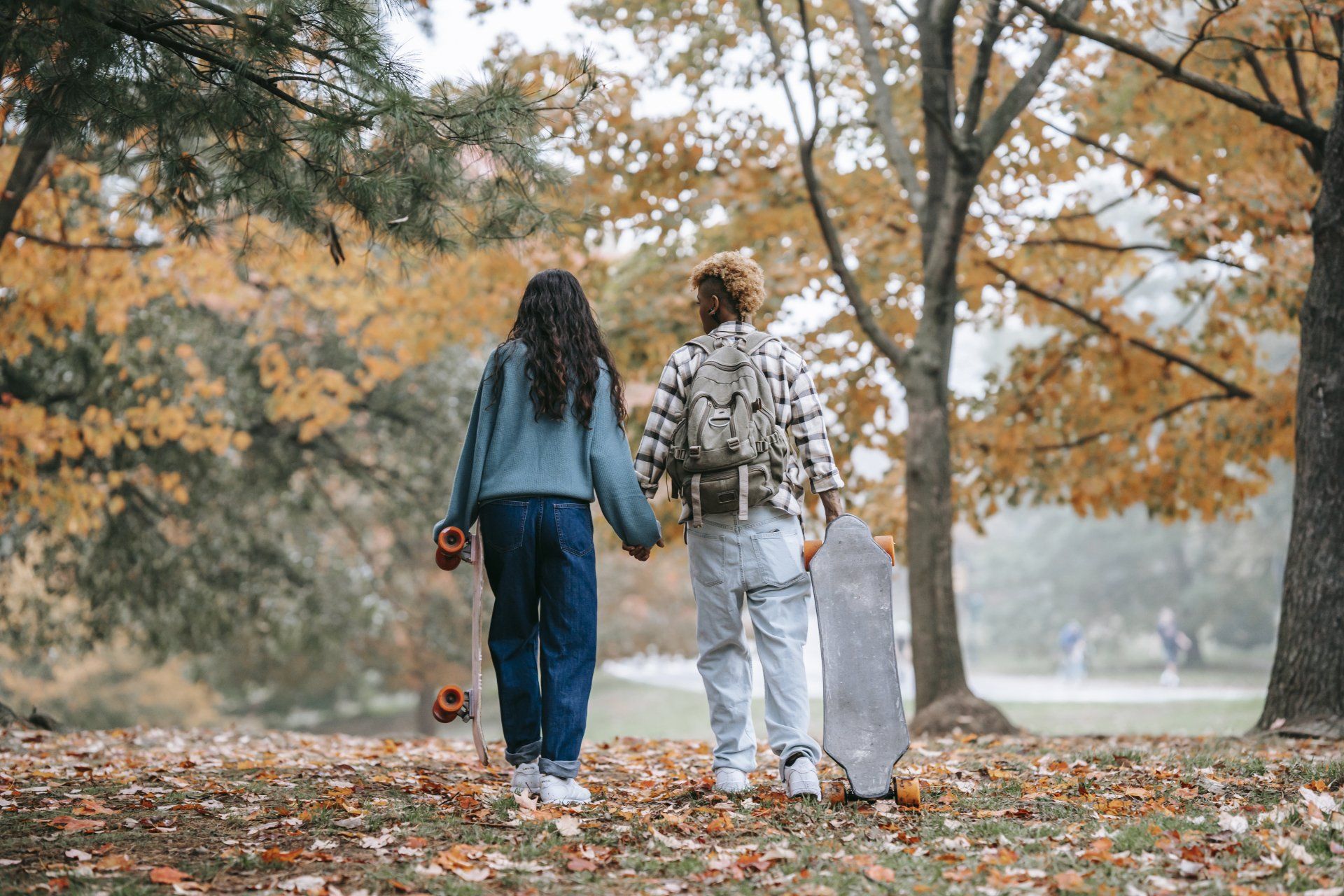 Couple holding hands, walking in a park, skateboards in hand. Fall foliage and trees in background.