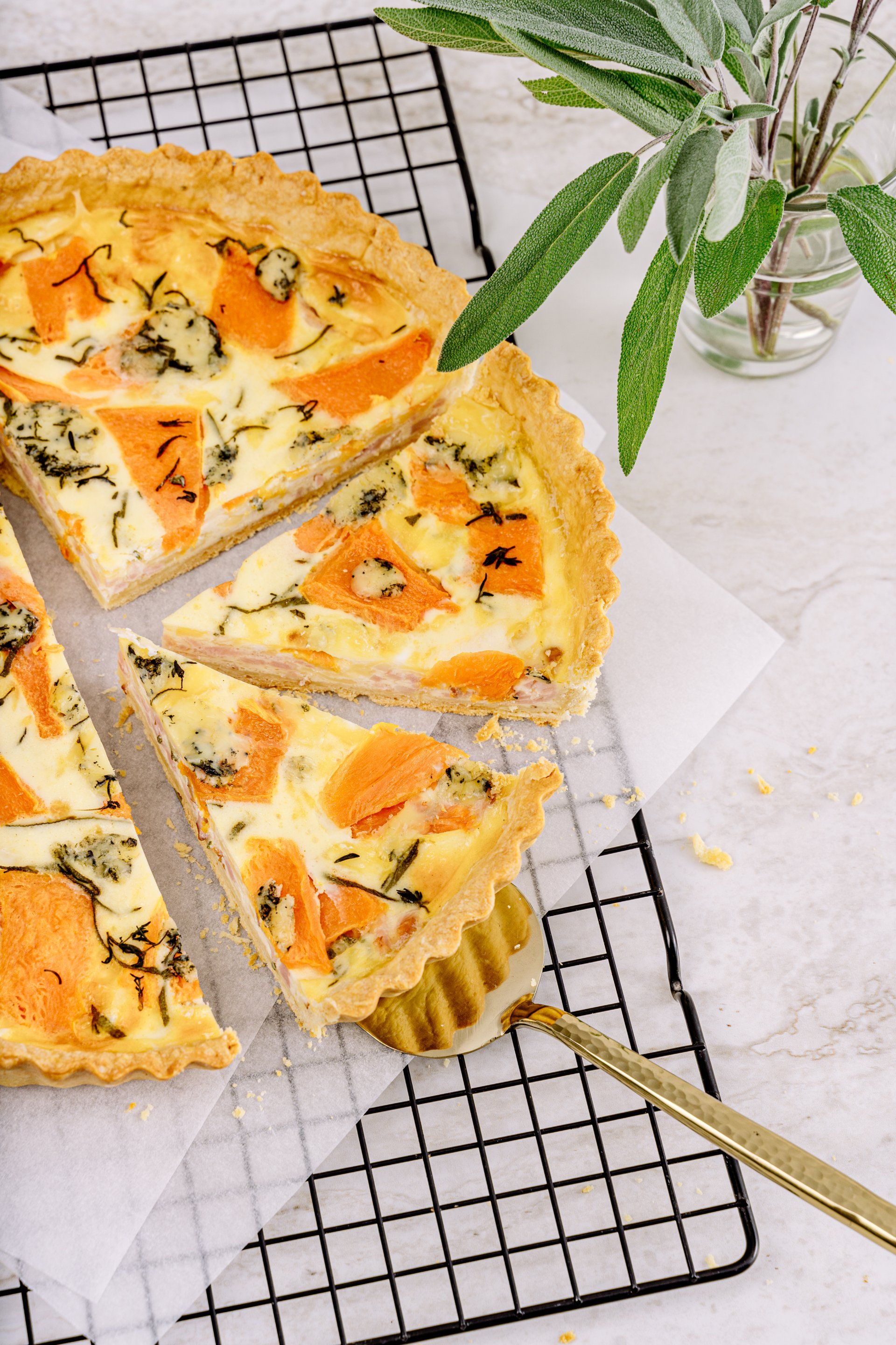 Sliced savory tart on a cooling rack with a gold pie server and a sprig of green leaves nearby