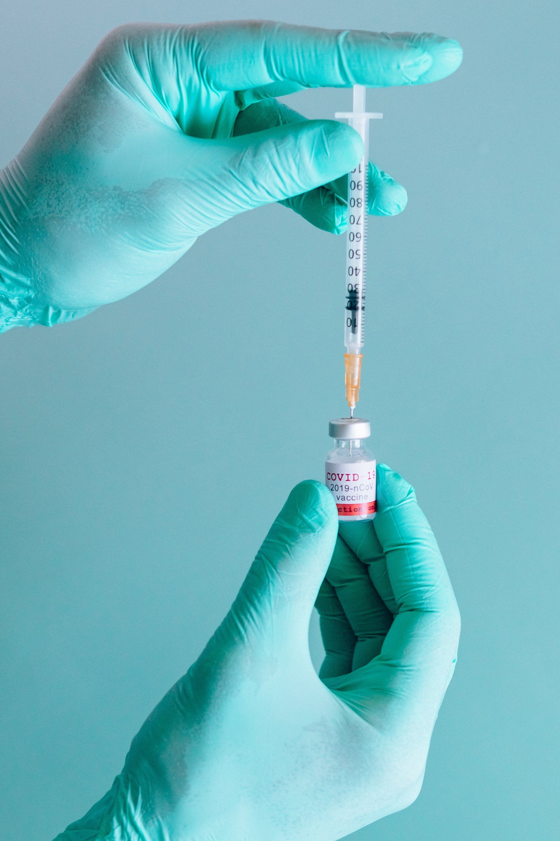 Hands in teal gloves drawing liquid from a vaccine vial with a syringe.