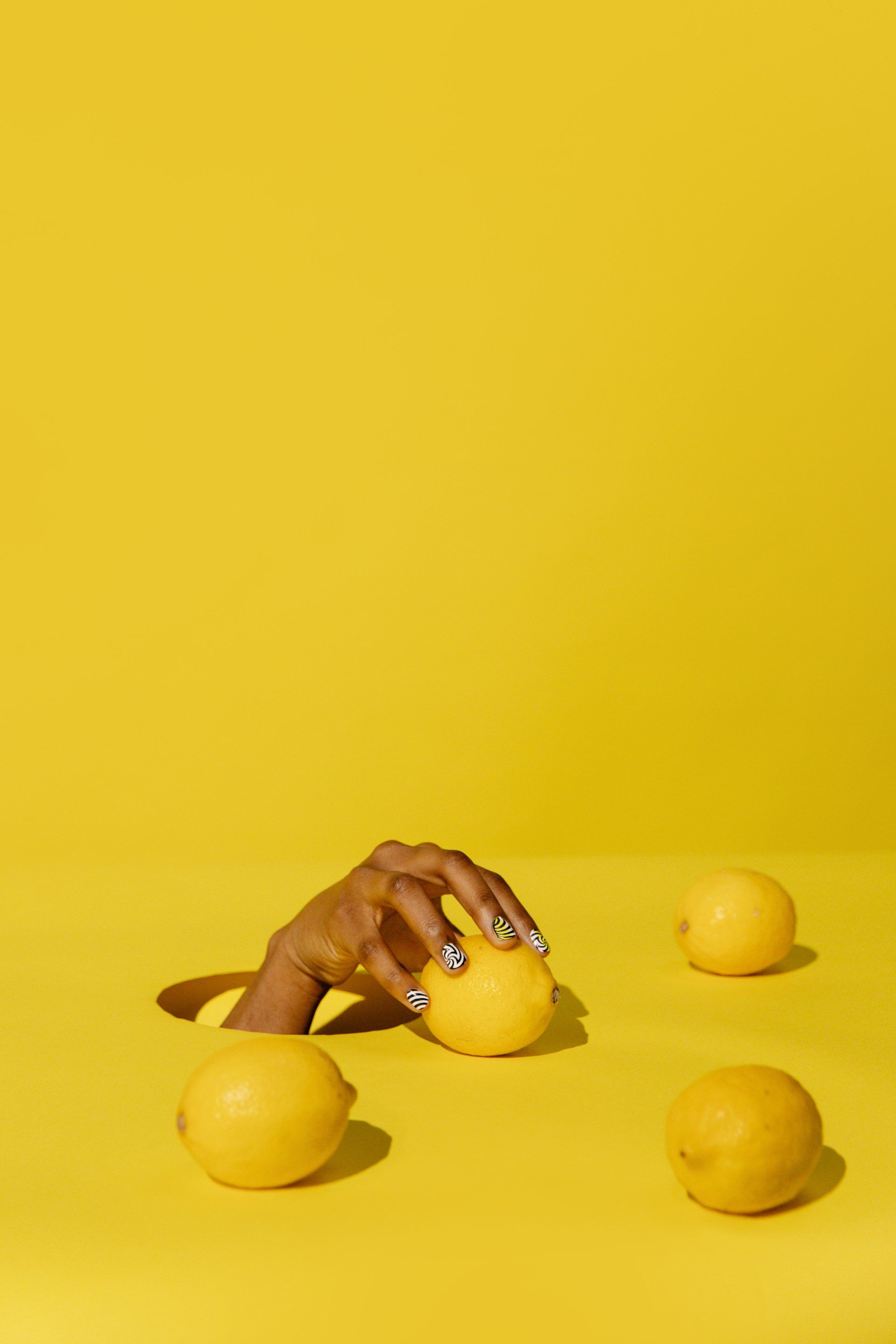 Hand reaching for a yellow lemon on a bright yellow background, with three other lemons nearby