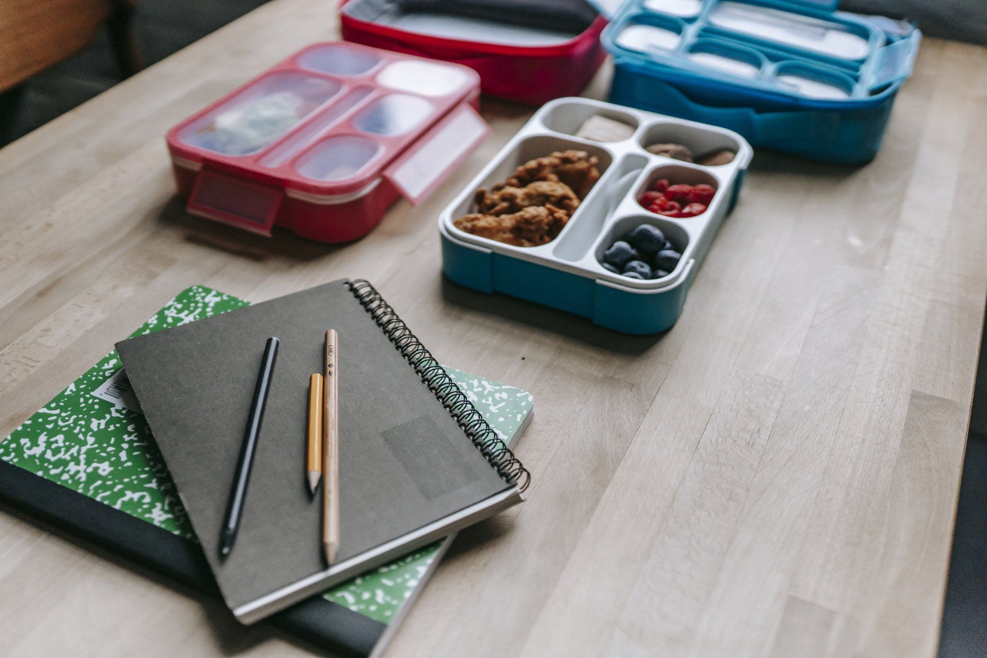 A wooden table topped with notebooks , pencils , and lunch boxes.