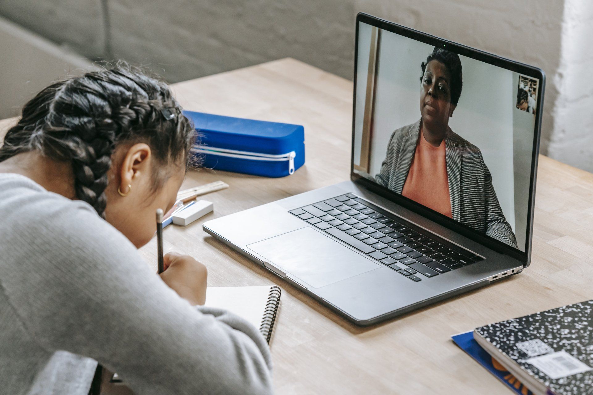 Person studying at a desk during a video call on a laptop, with notebooks and a pen nearby