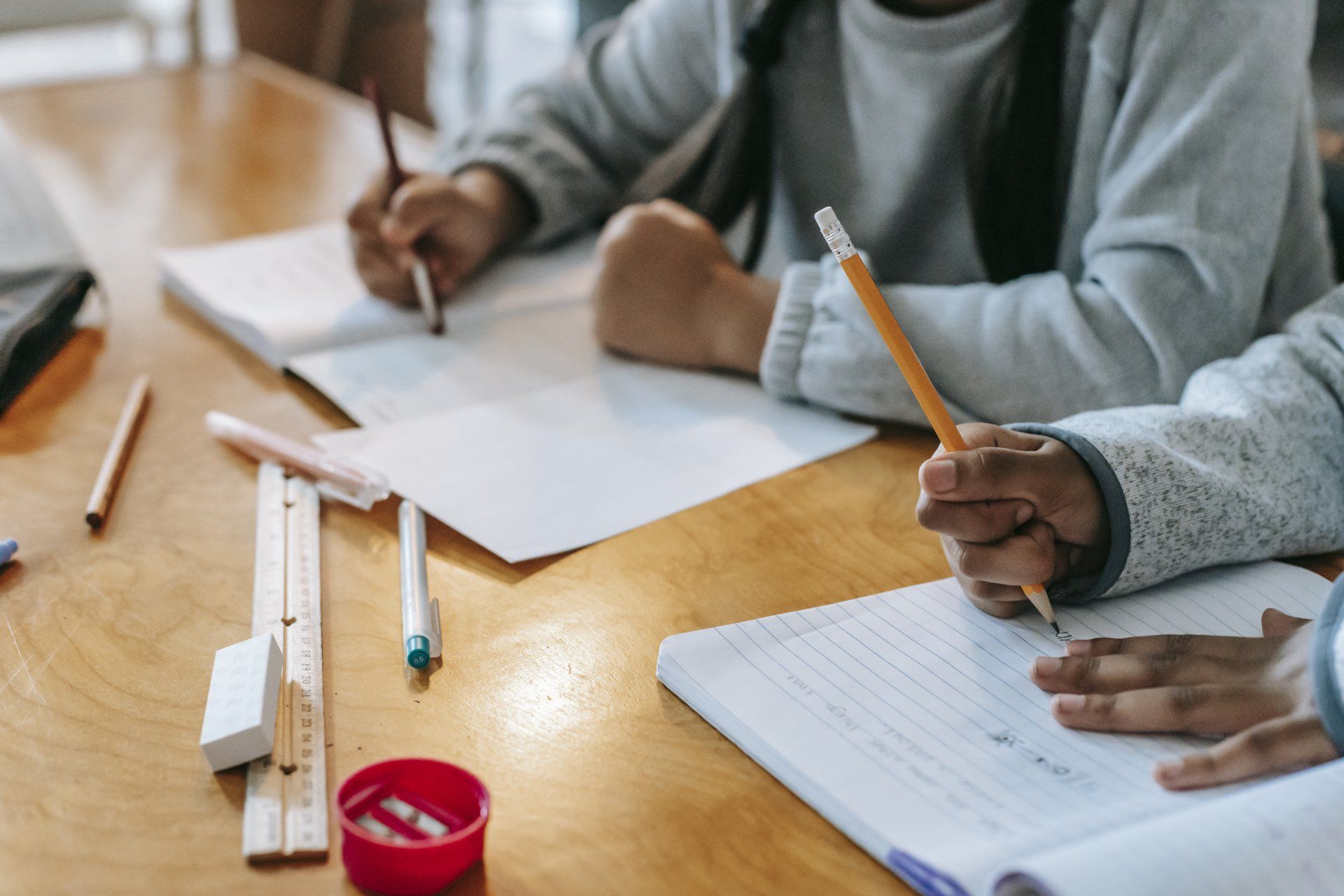 Twee kinderen schrijven in schriftjes aan een houten tafel met potloden en schoolspullen.