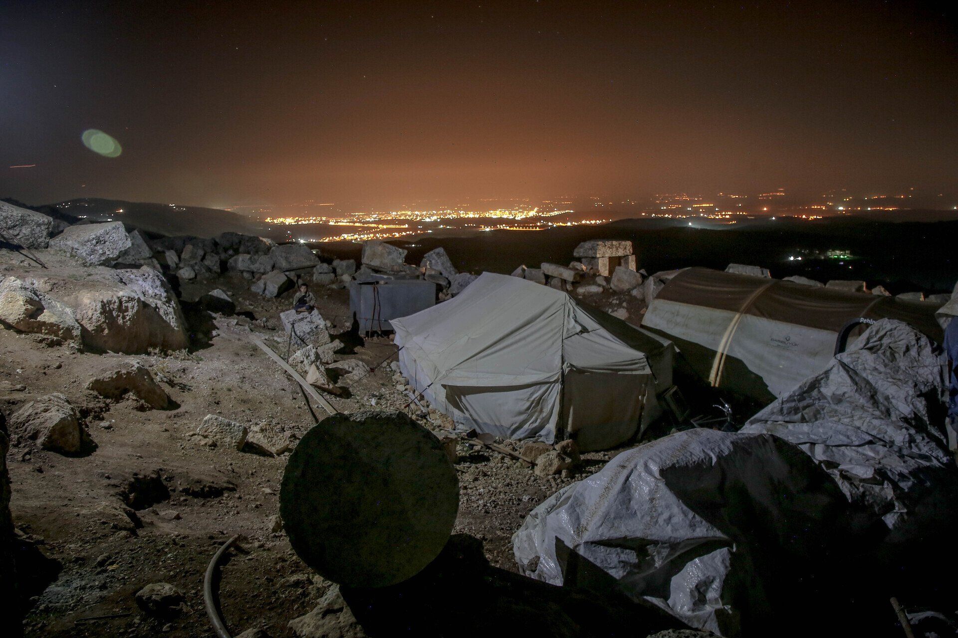 Nighttime camp with tents on rocky terrain overlooking distant city lights