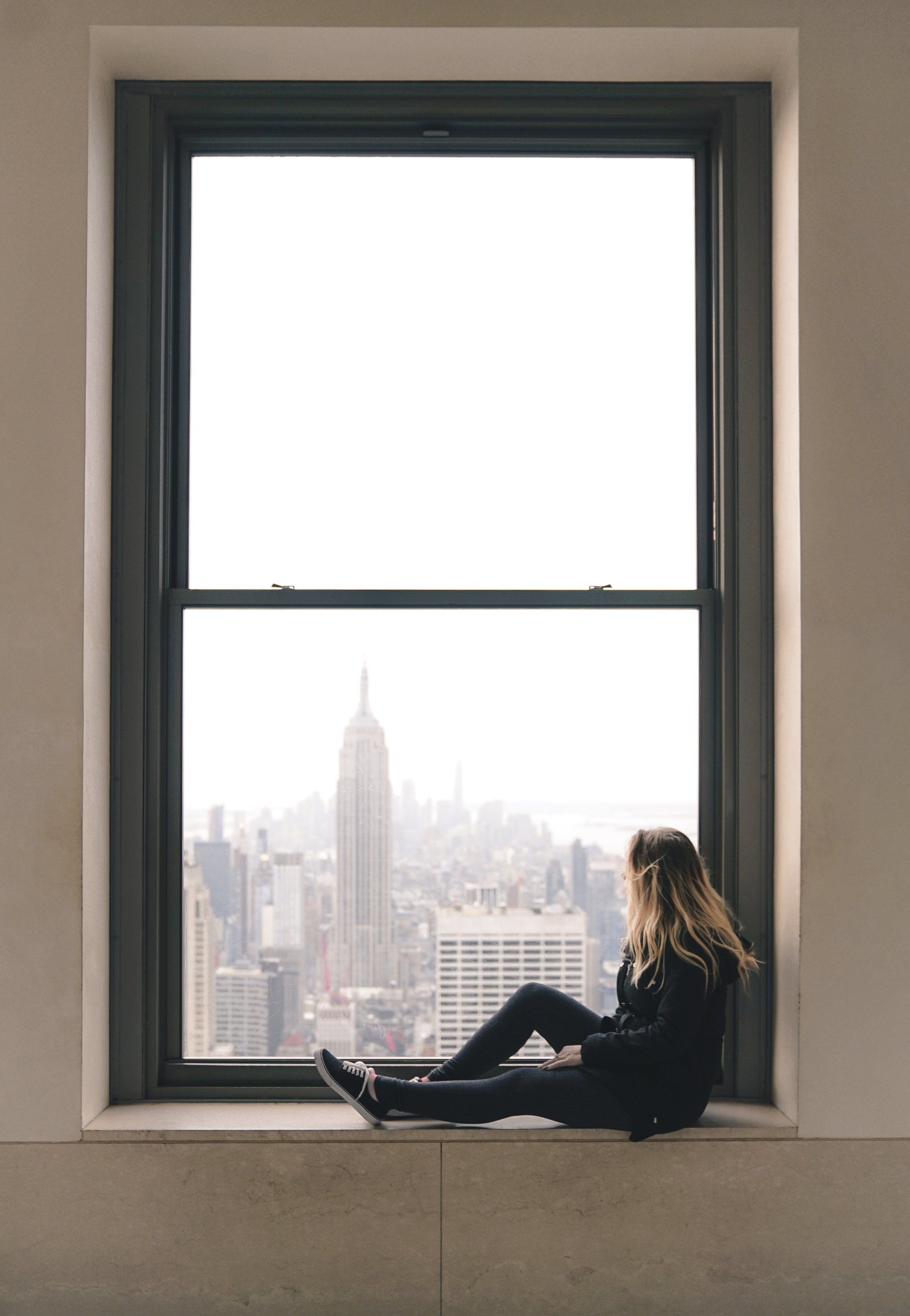 A woman is sitting on a window sill looking out at the city.