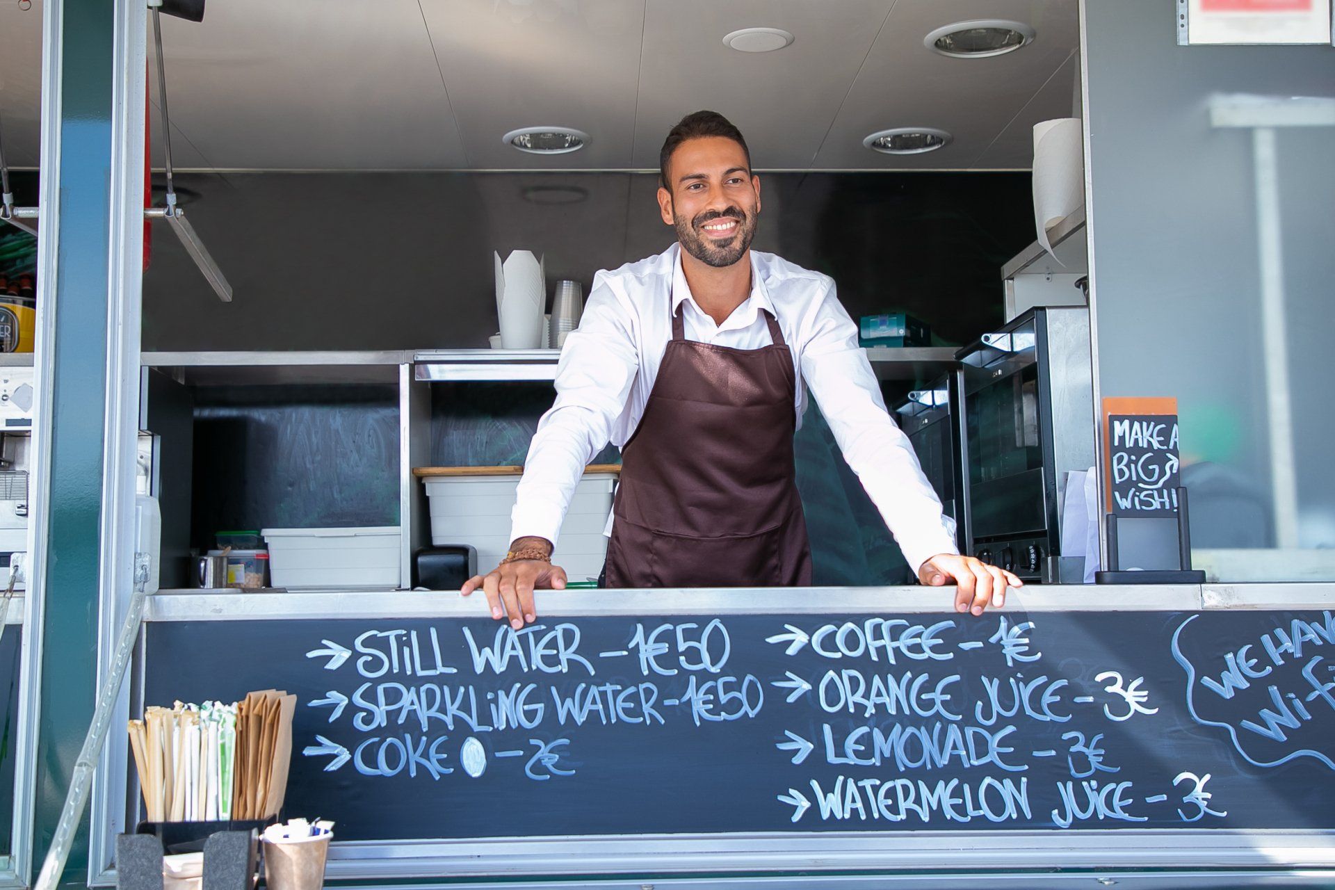 Man in apron at food truck window smiles, drink prices on chalkboard: coffee, water, juice.