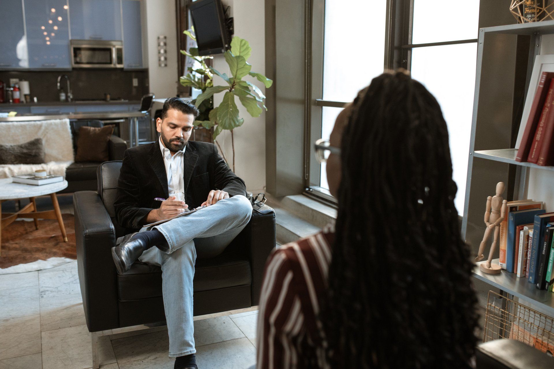Two people seated in a living room, having a conversation near a bookcase and large windows.