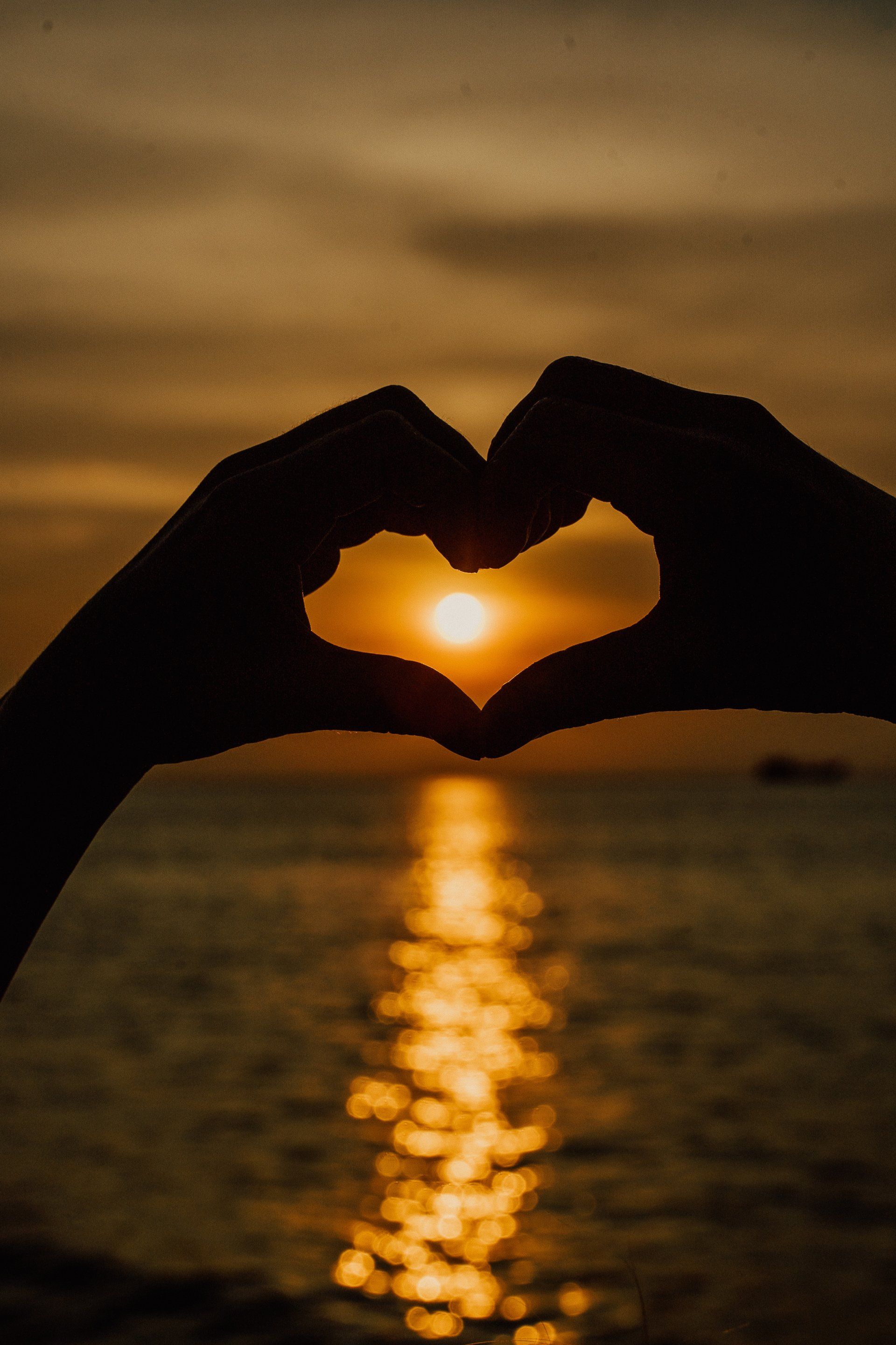 Hands making a heart shape around the setting sun over the ocean.