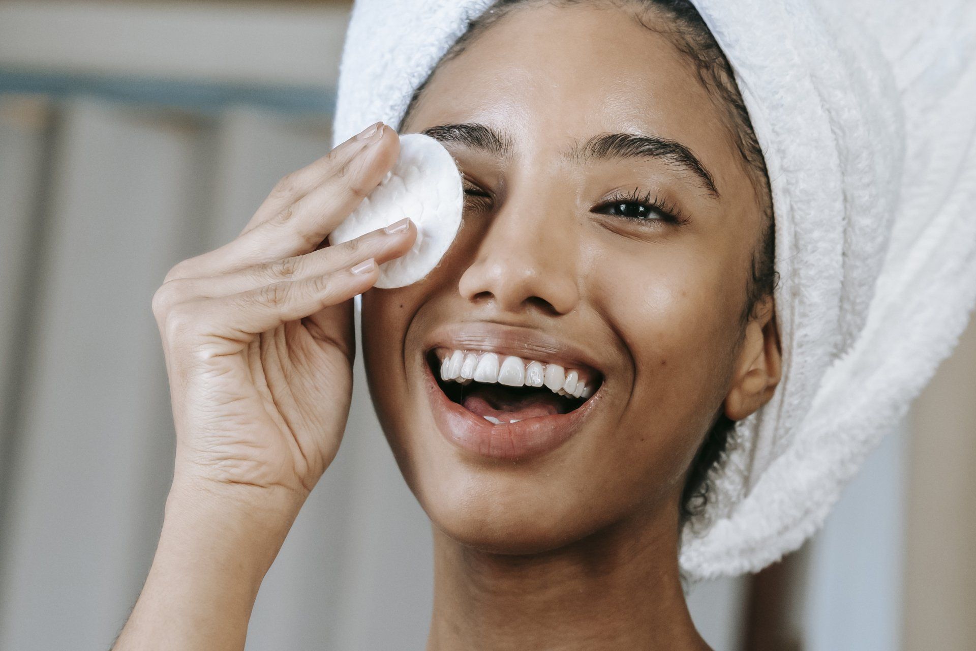 A woman with a towel wrapped around her head is cleaning her face with a cotton pad.