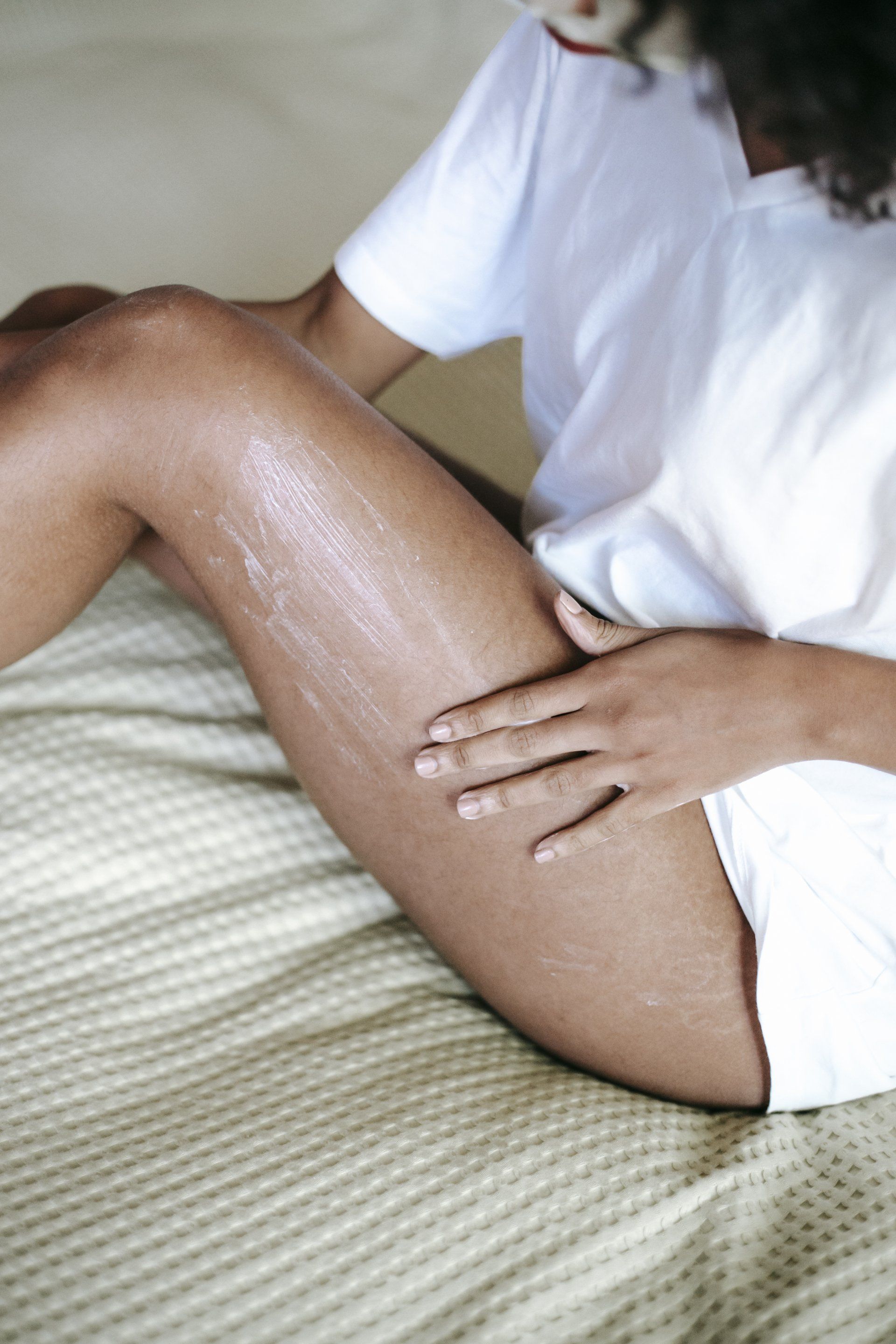A woman is sitting on a bed applying lotion to her leg.