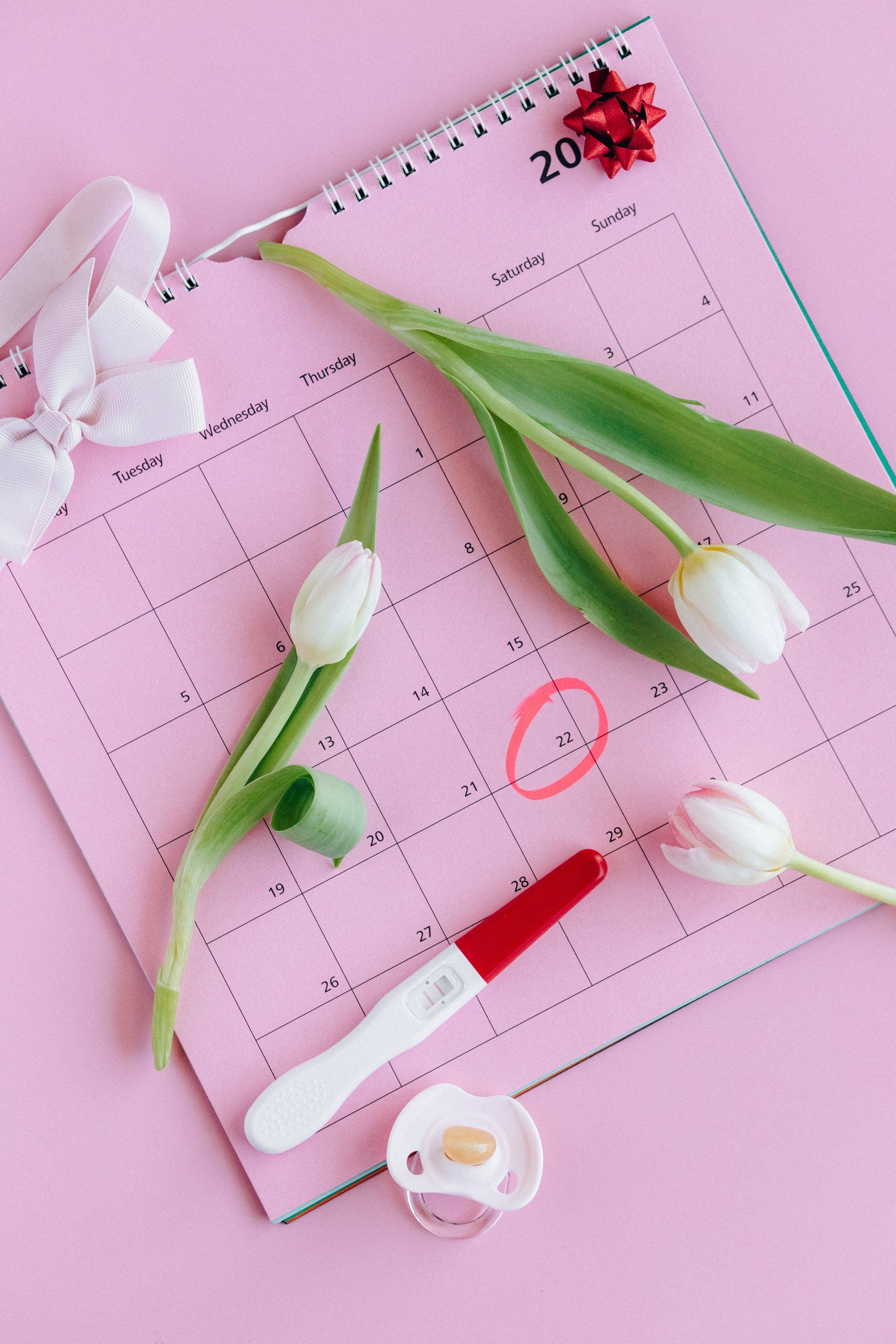 Pink calendar with a red circle, pregnancy test, white tulips, and a pacifier.