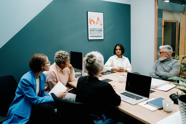 People in a meeting room, sitting around a table with laptops, notebooks, and a framed print on the wall.