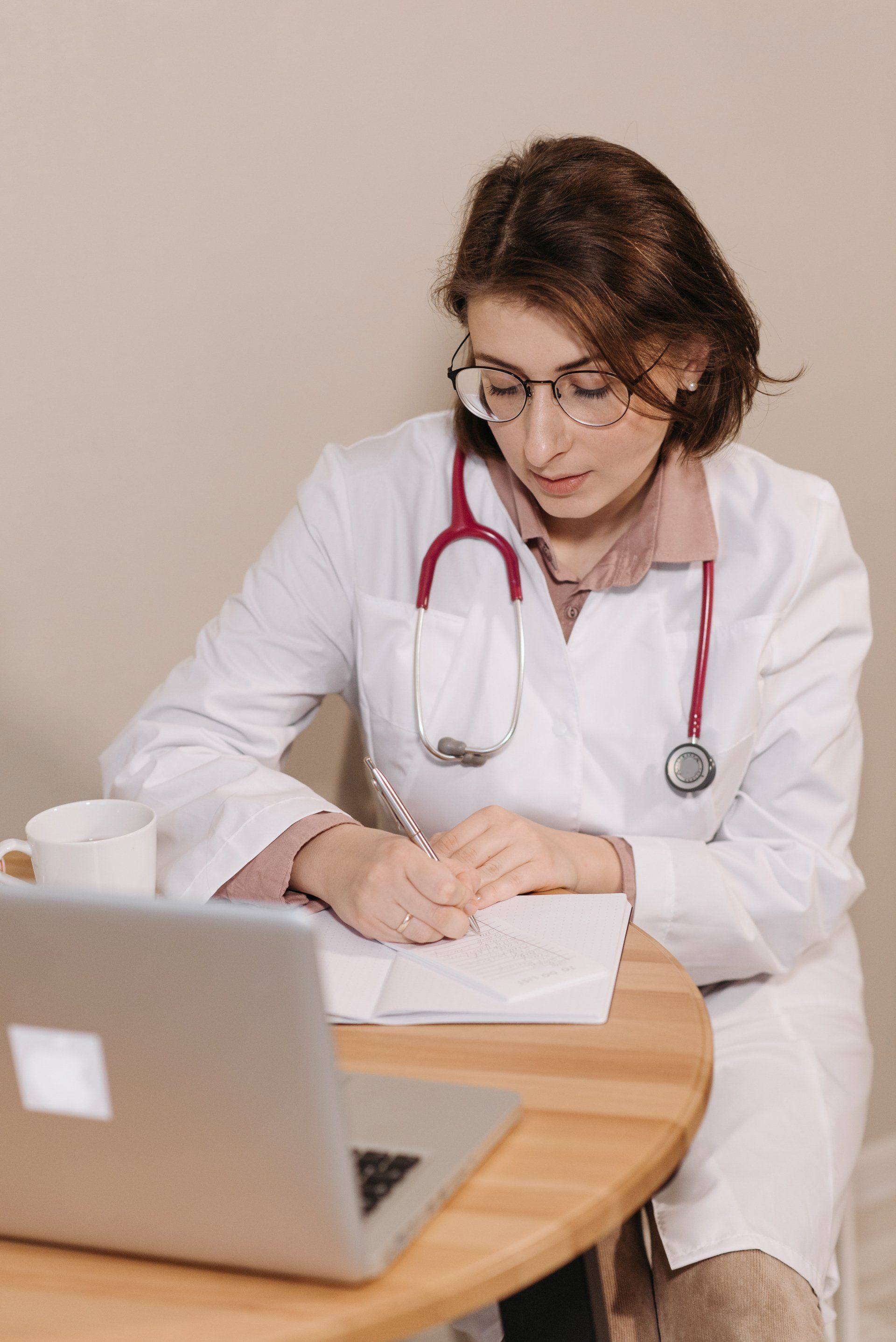 A medical professional in a white coat and stethoscope writing on a notepad while seated at a desk with a laptop.