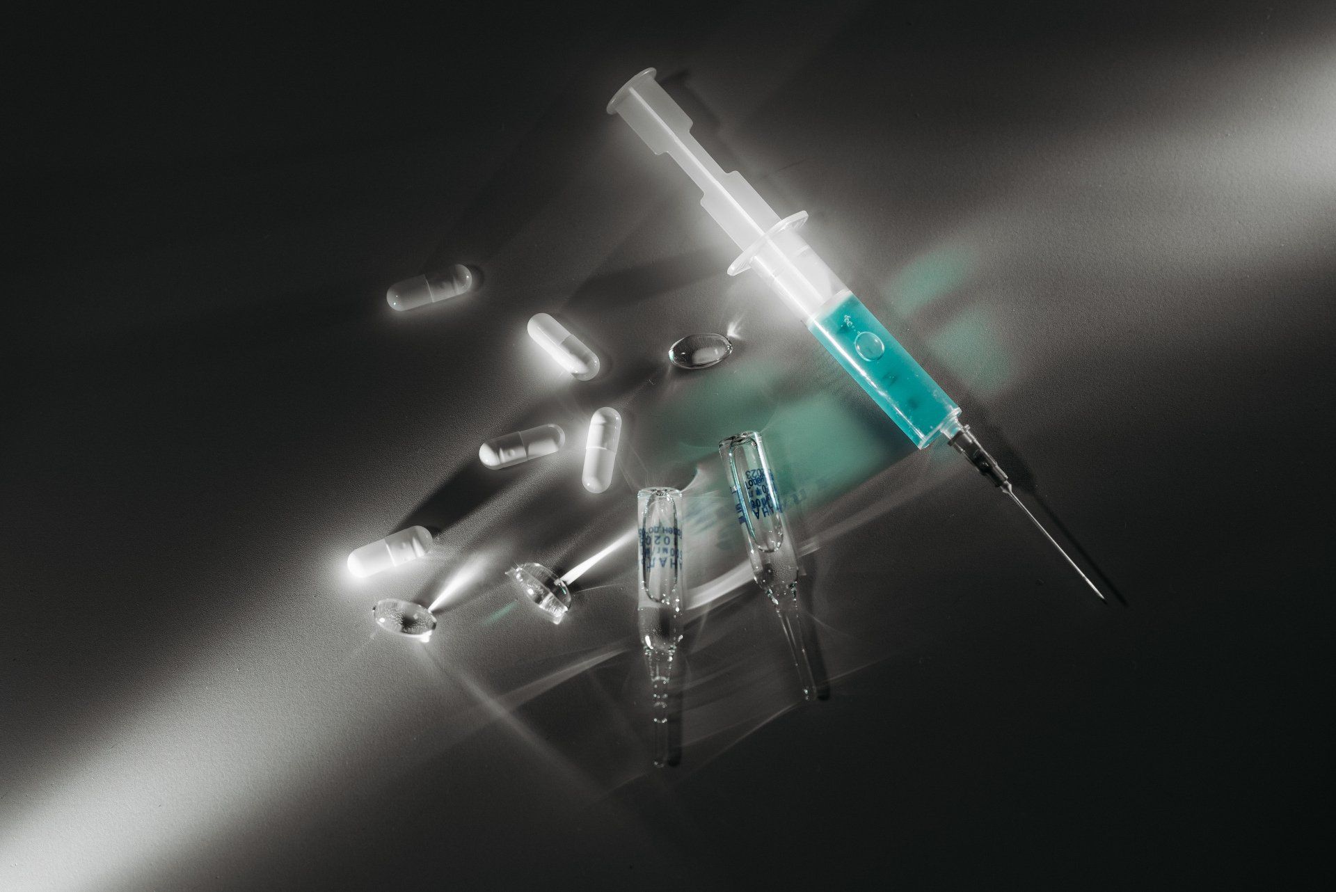 Syringe with blue liquid, pills, and vials on a dark reflective surface.
