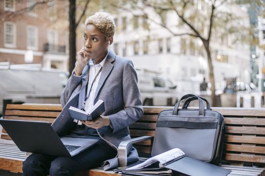 A woman is sitting on a bench with a laptop and a briefcase.