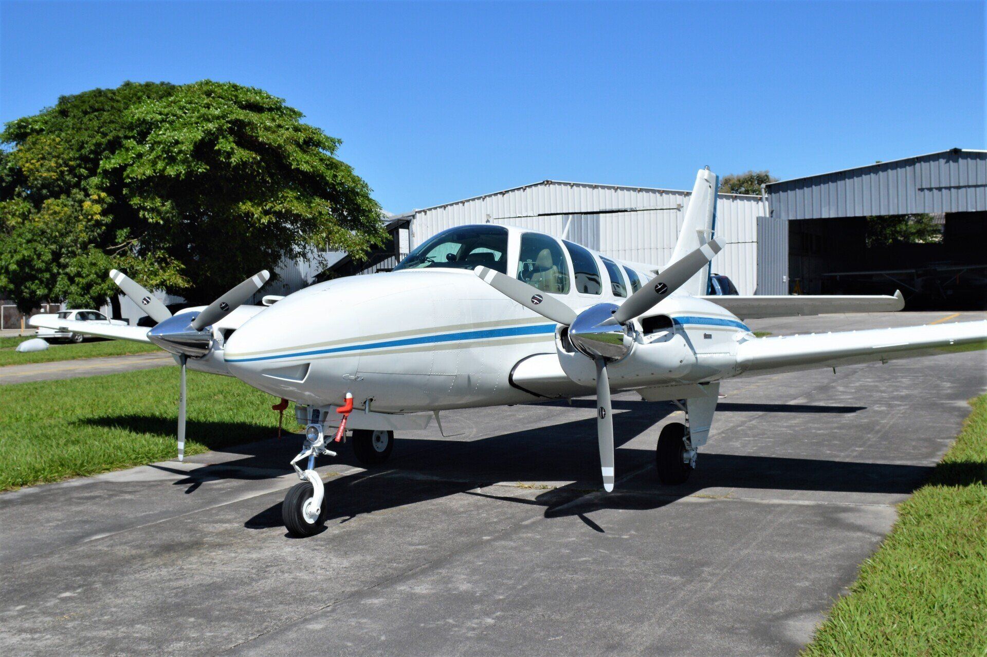 White twin-engine propeller plane parked on a driveway beside a hangar in bright sunlight