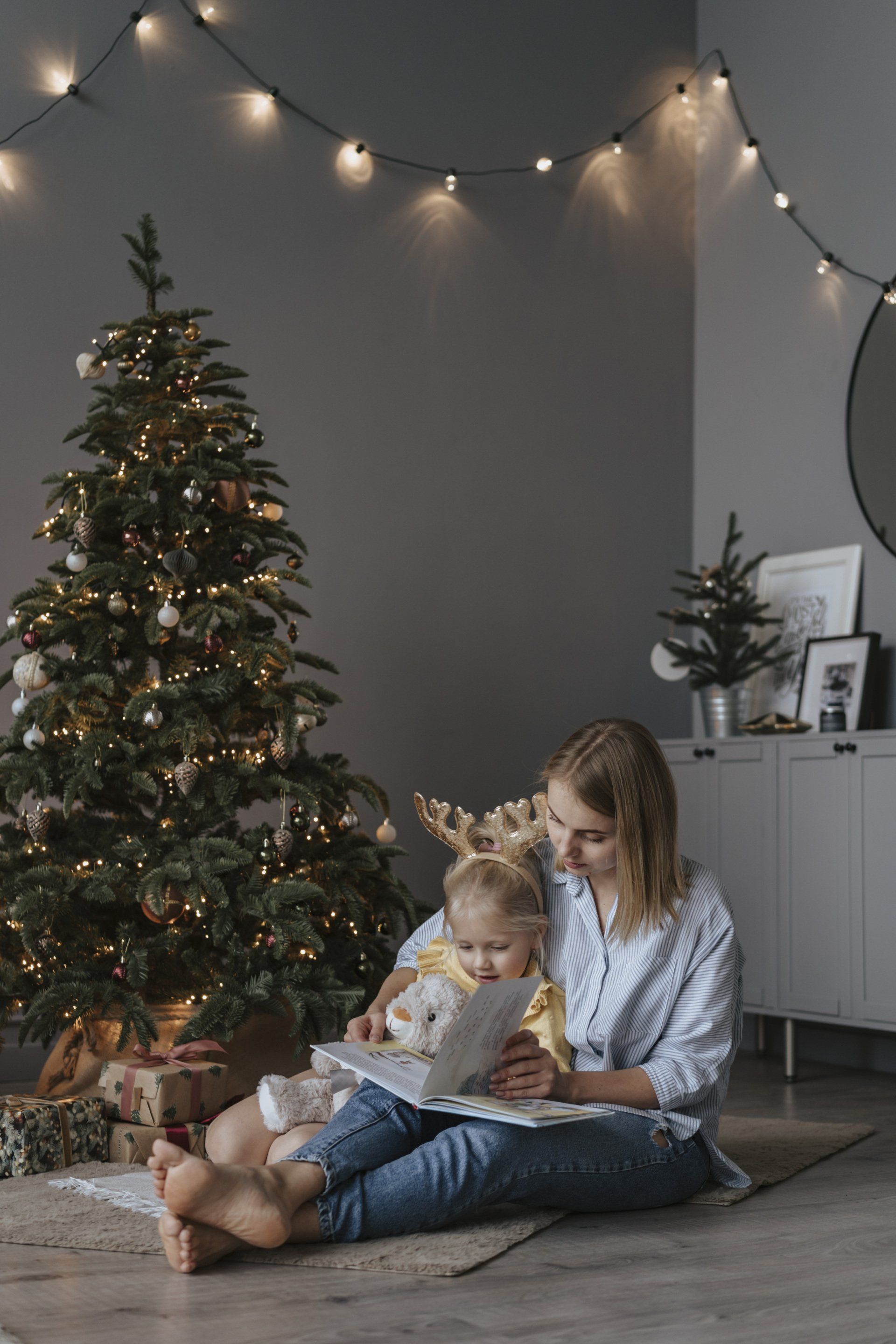 A woman is sitting on the floor reading a book to a baby in front of a christmas tree.