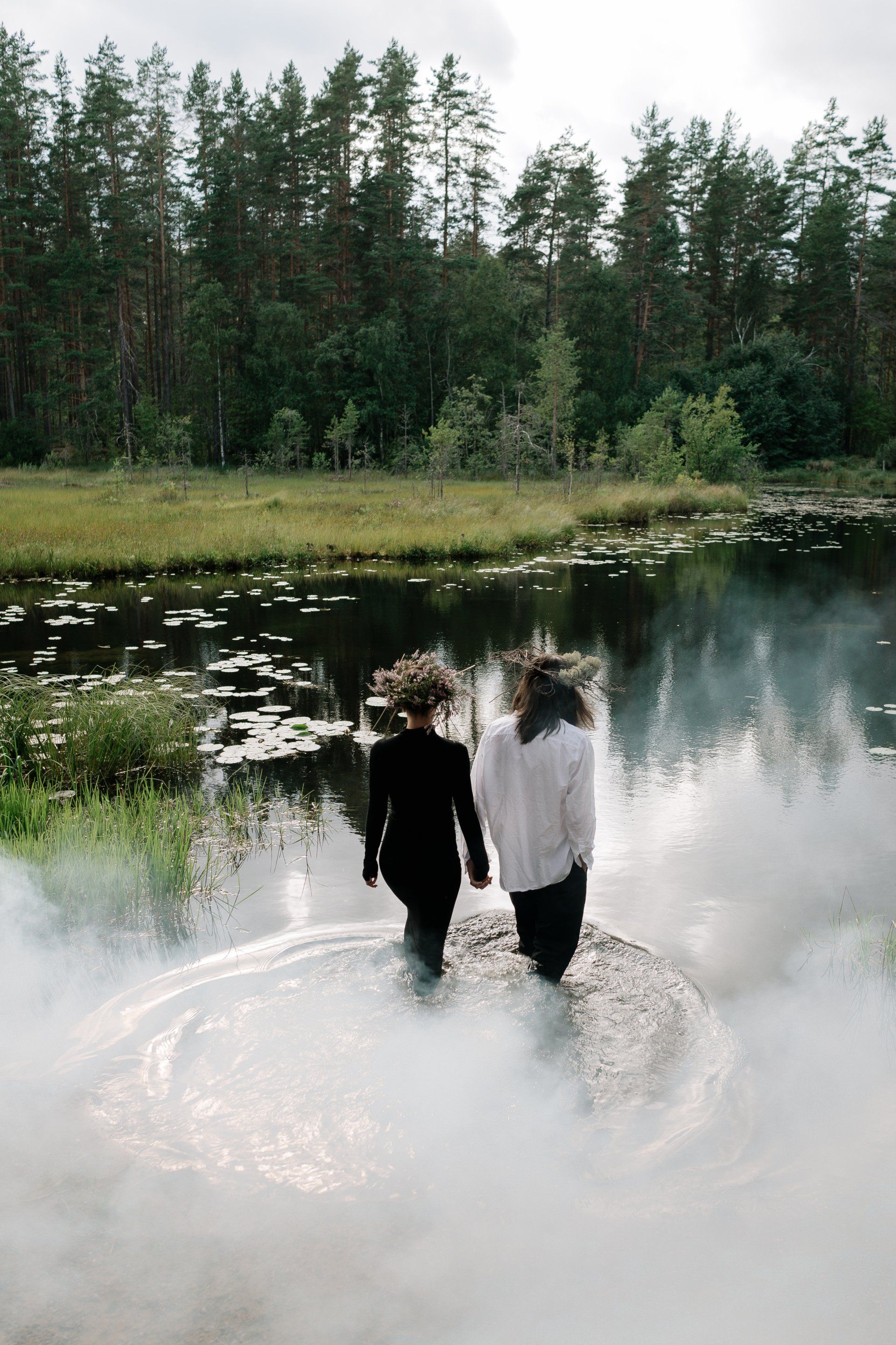 Two people walk into water, surrounded by smoke, a forest and lake in background.