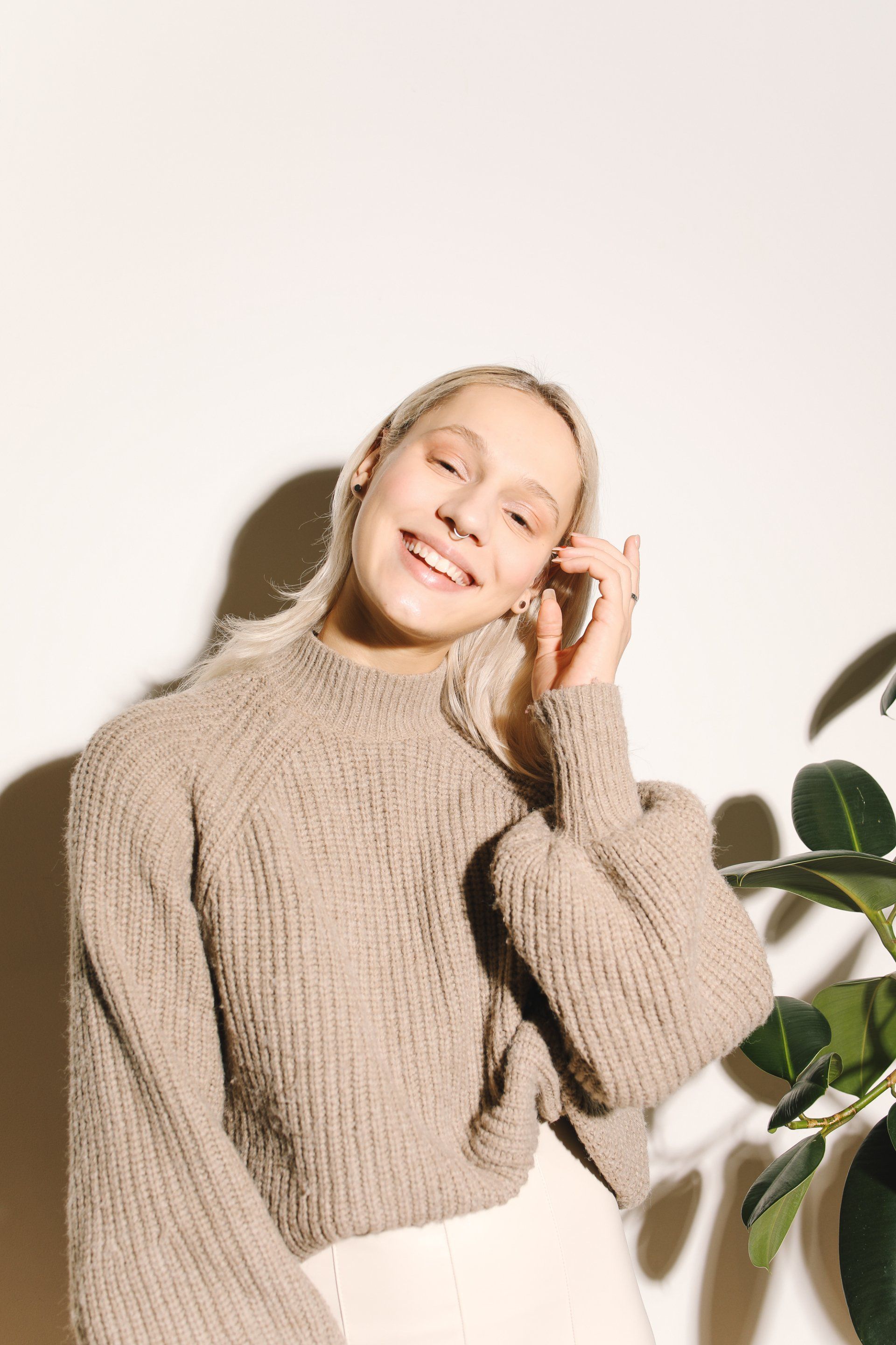 Vrouw in beige trui glimlacht, hand in haar haar, vlakbij een plant.