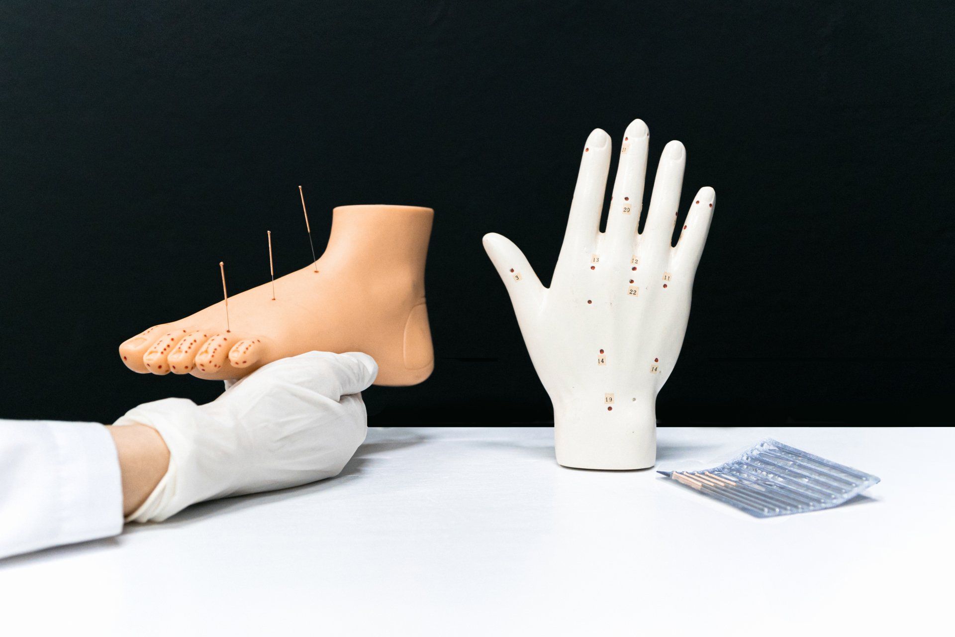 Person holding prosthetic foot, with acupuncture needles, next to a prosthetic hand, both on a white table.