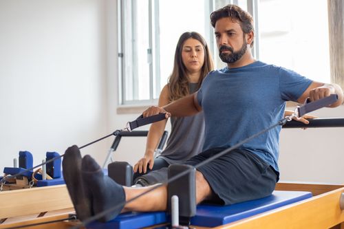 Man using a Pilates reformer with a trainer assisting in a bright studio