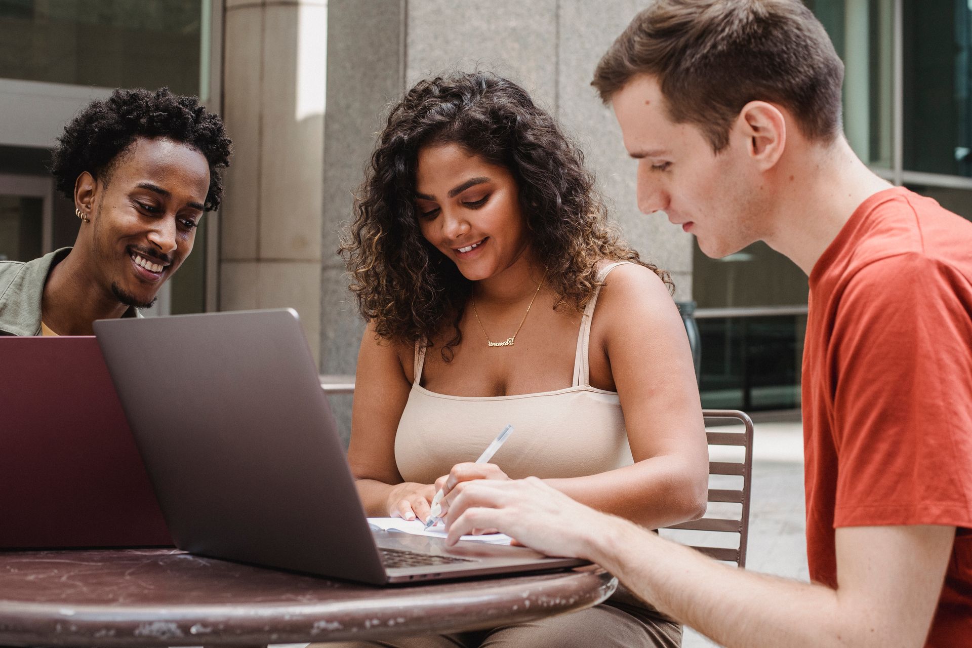 Three people gathered around a laptop at an outdoor table, smiling and looking at the screen