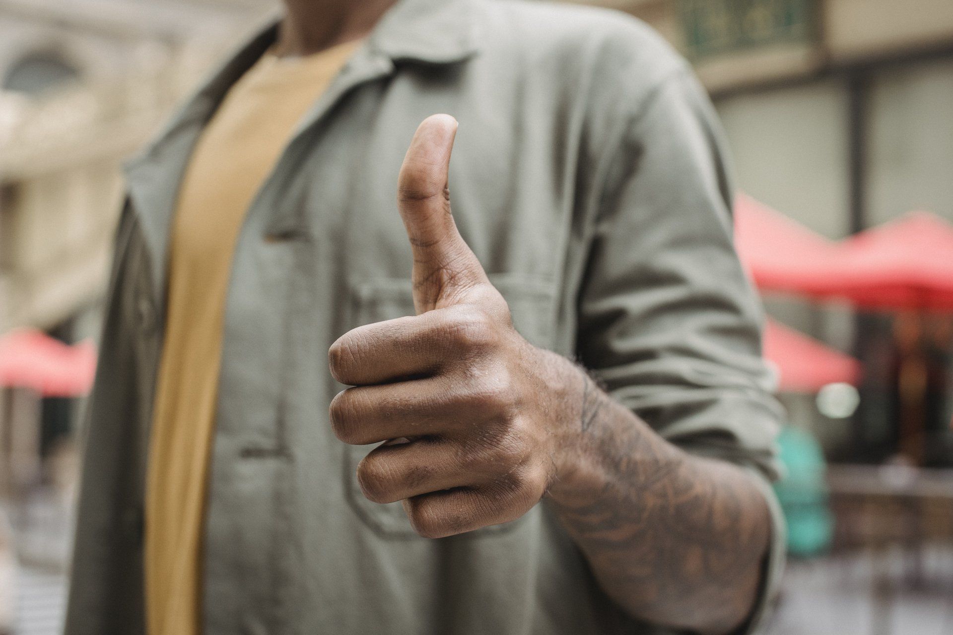 Person in olive jacket gives a thumbs-up gesture outdoors, orange shirt visible, blurred background.