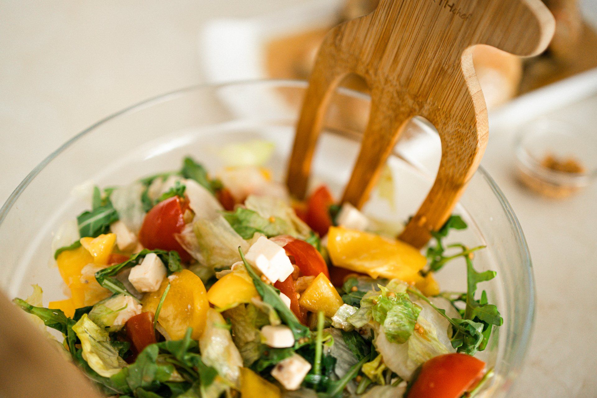 Salad in a glass bowl with wooden tongs, including tomatoes, peppers, and feta cheese.