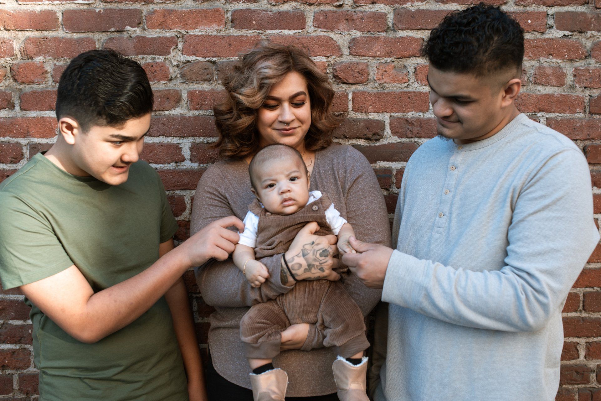 A woman holds a baby, flanked by two people, standing against a brick wall.