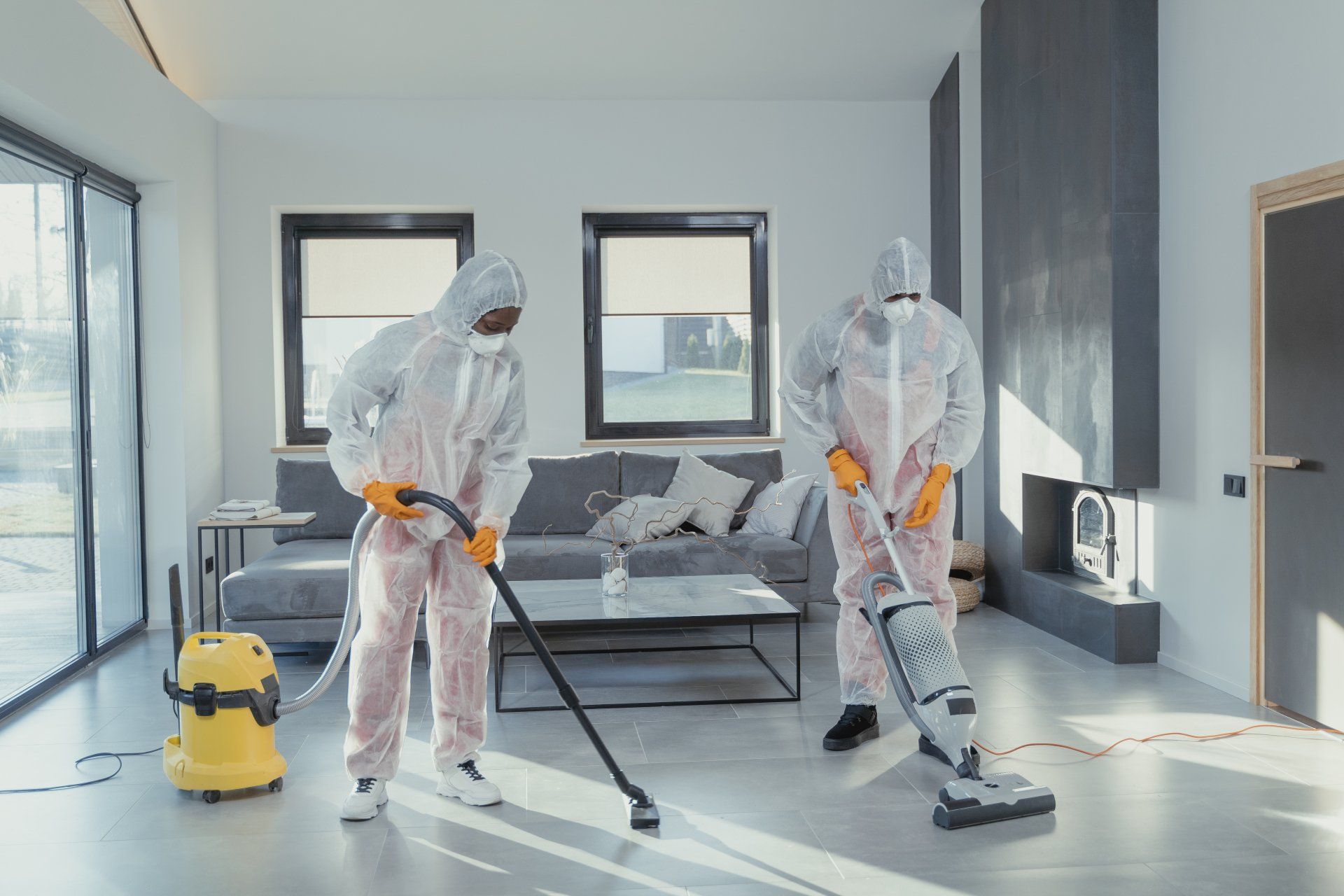 Two people in hazmat suits vacuuming a modern living room. One uses a hose, the other a vertical vacuum.