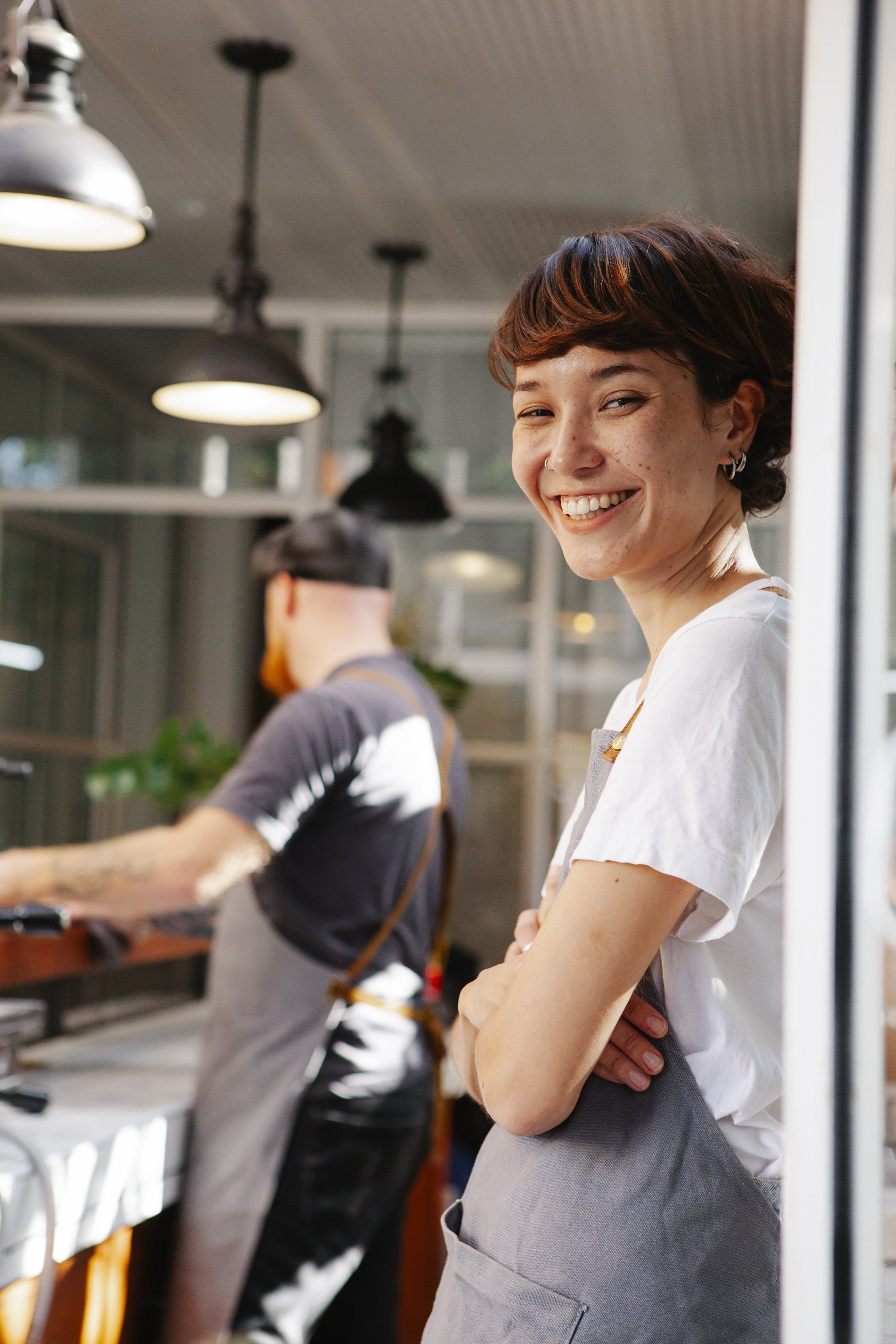 A smiling person in a white t-shirt and grey apron stands in a cafe with a colleague working in the blurred background.