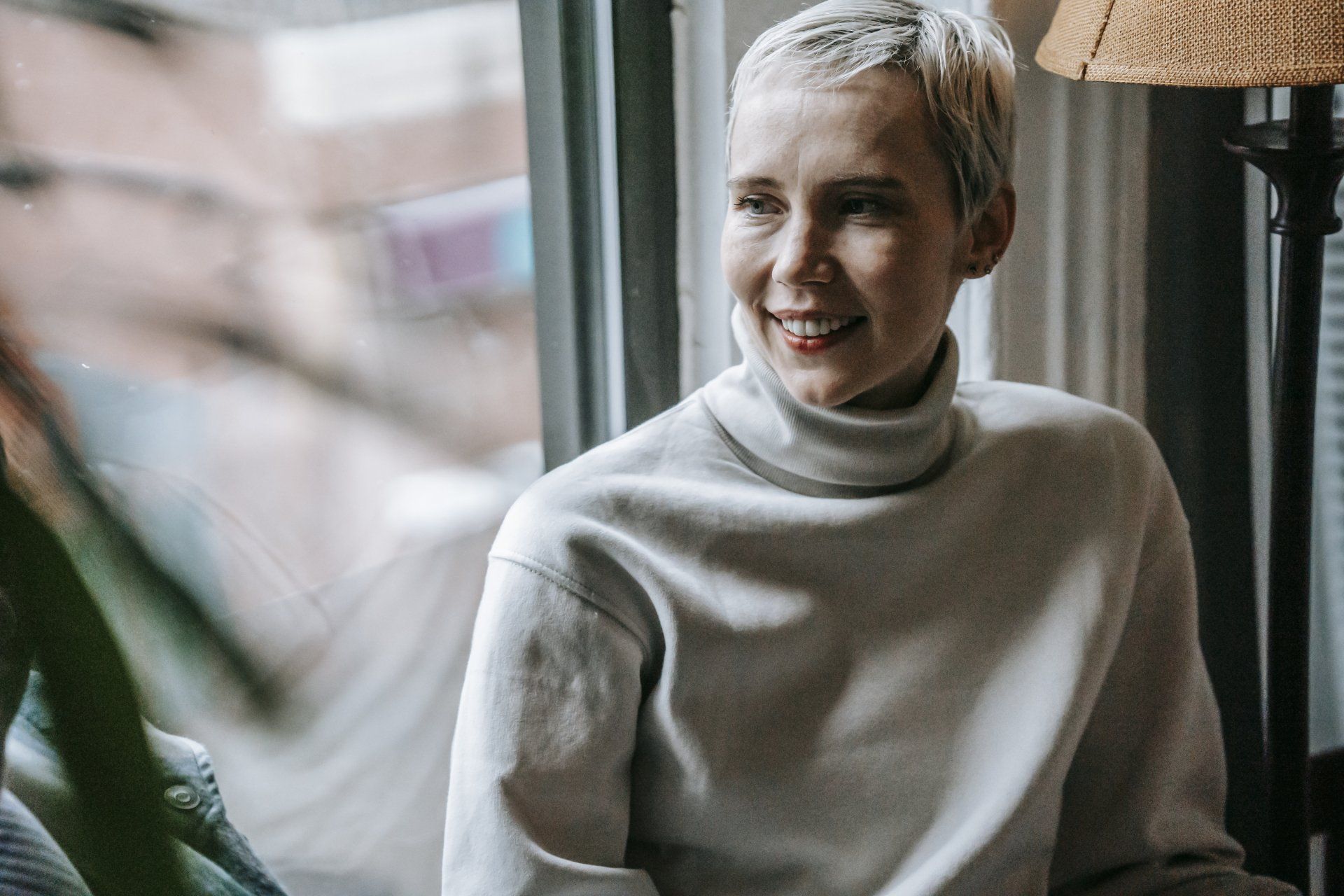 A woman is sitting in front of a window wearing a turtleneck sweater and smiling.