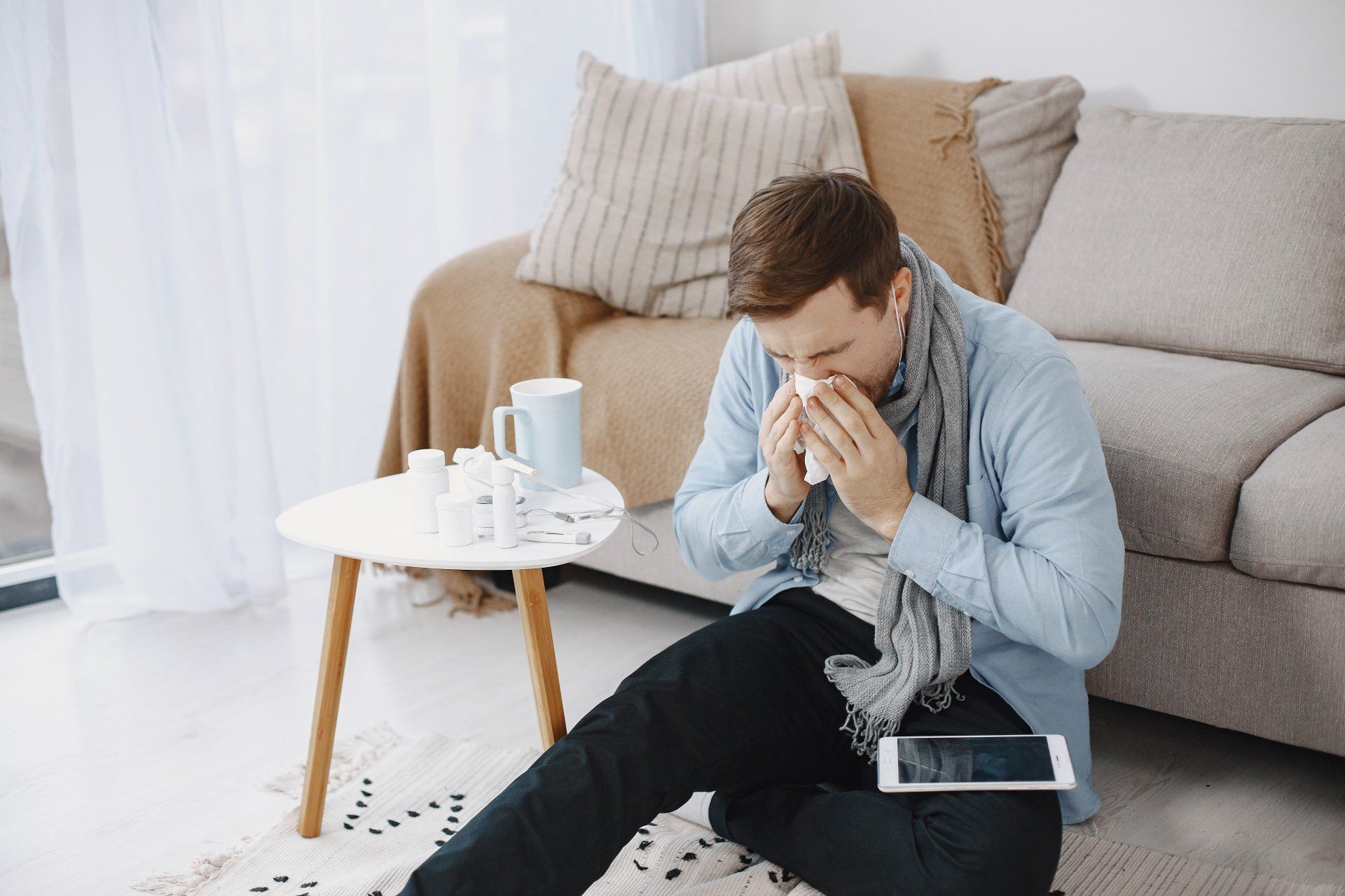 Man sitting on the floor, blowing nose, with medicine and a tablet nearby.