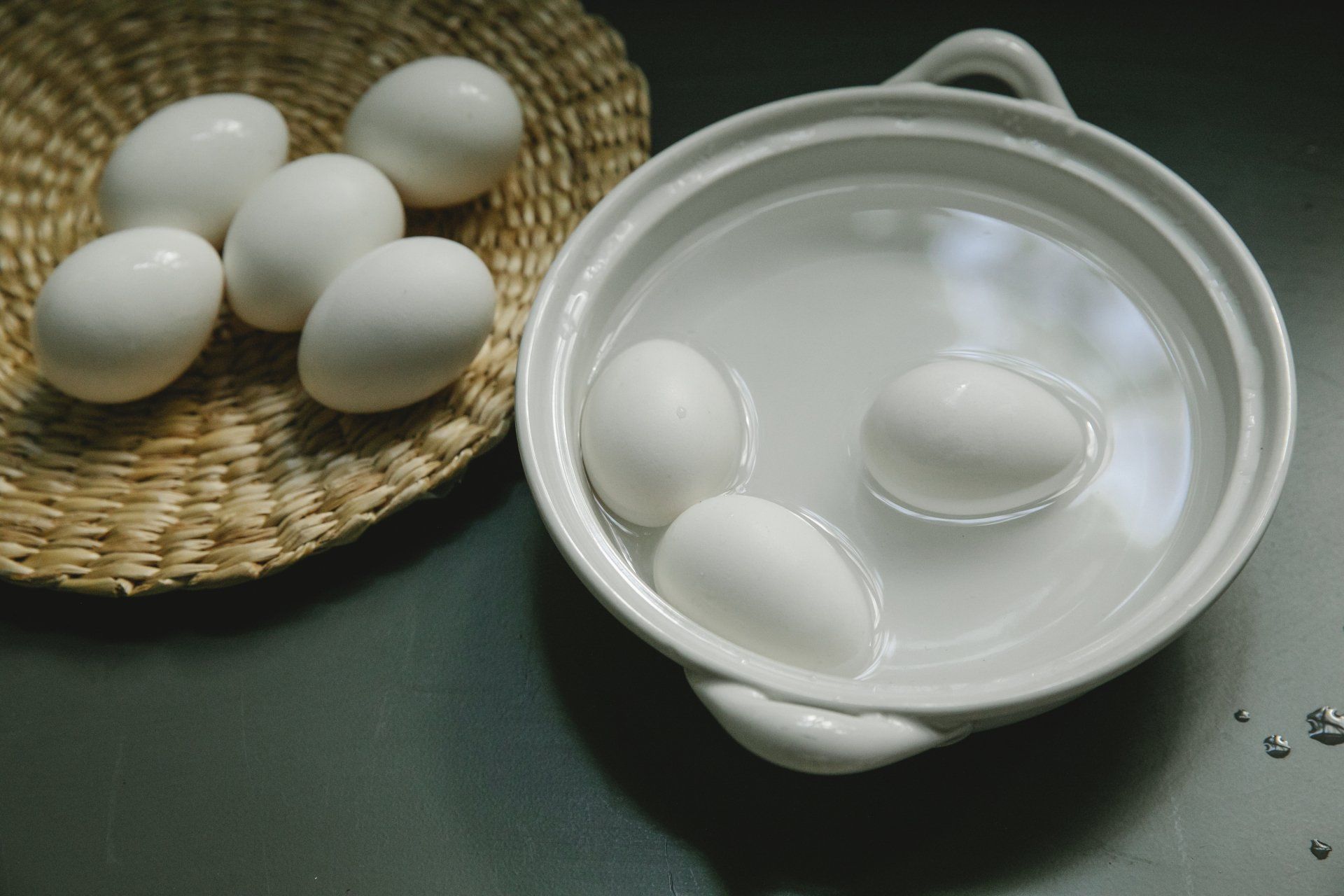 White eggs in a bowl of water and in a woven basket on a dark surface.