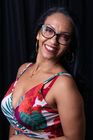 Smiling person wearing glasses, hoop earrings, and a floral dress against a black background.