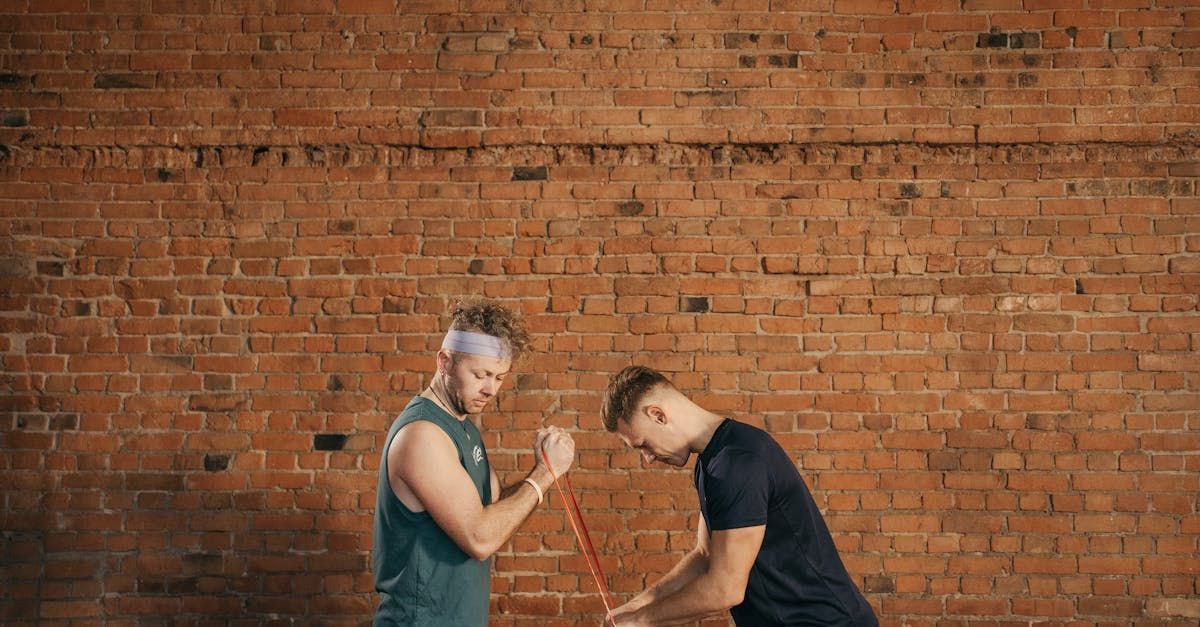 Two people exercising with a red bar in front of a brick wall. One person watches while the other bends over.