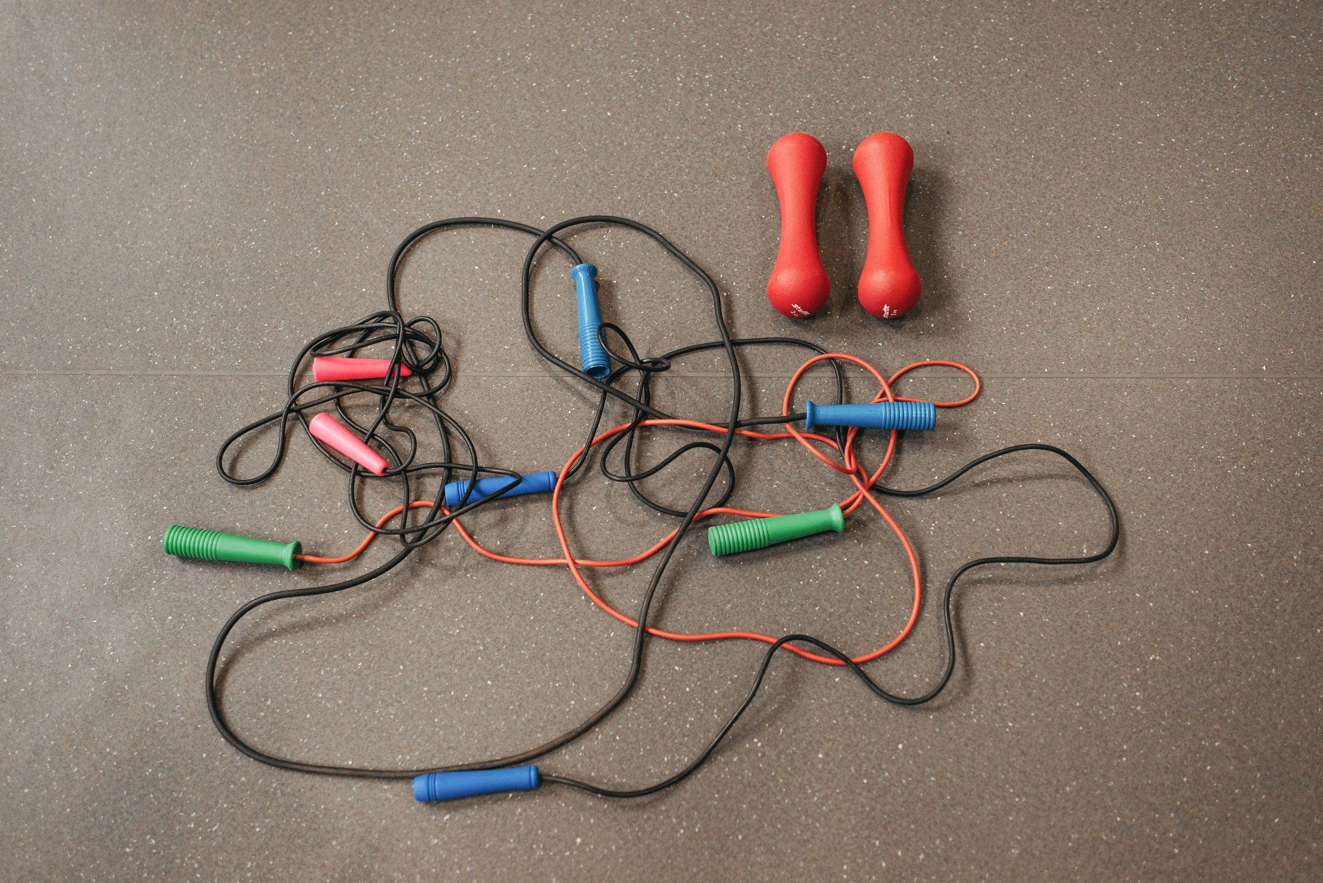 Several jump ropes with colored handles and two red hand grippers on a gray surface