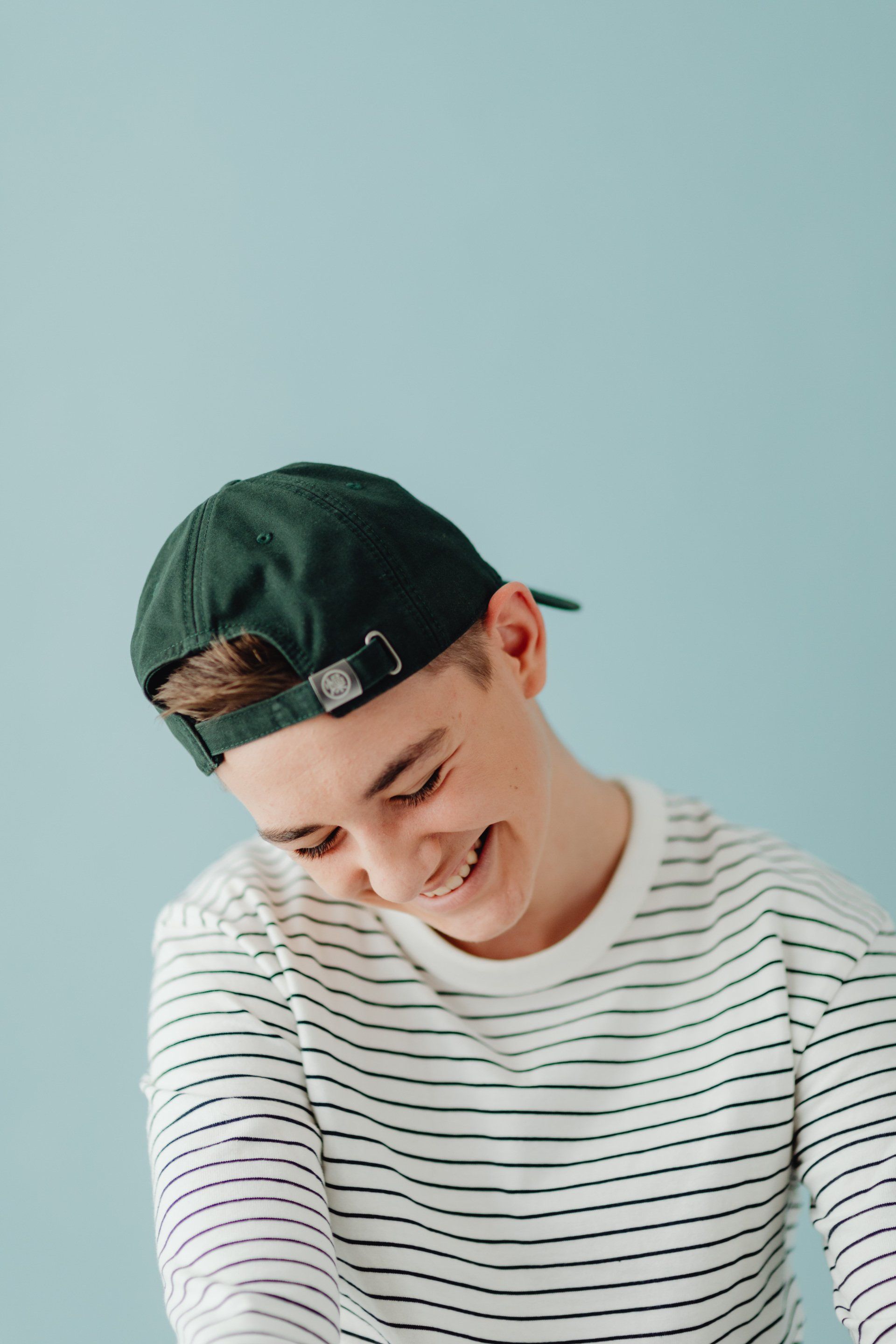 A young man wearing a striped shirt and a green baseball cap is smiling.