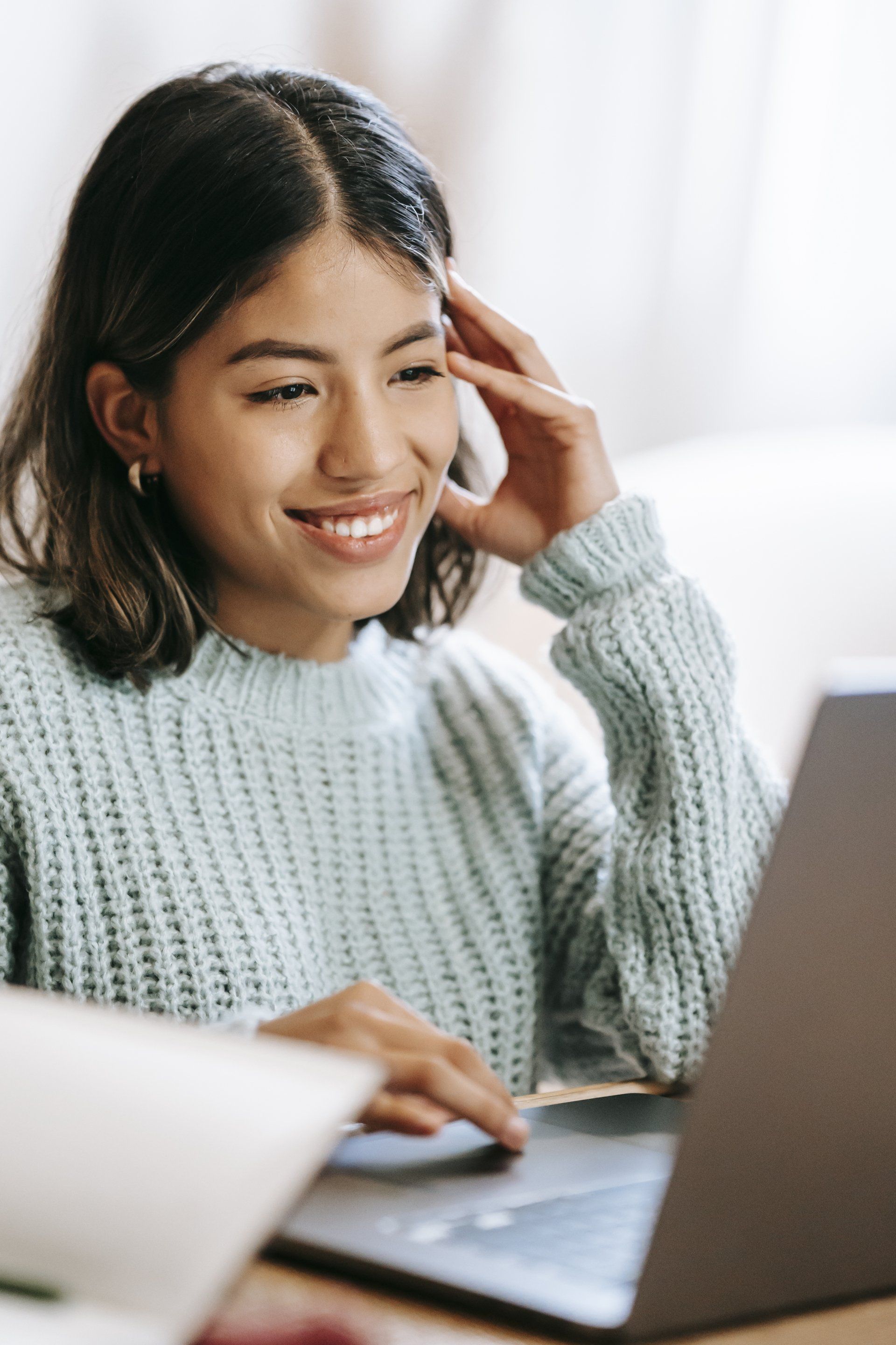 Woman smiling at a laptop, hand on her head, wearing a light blue sweater.